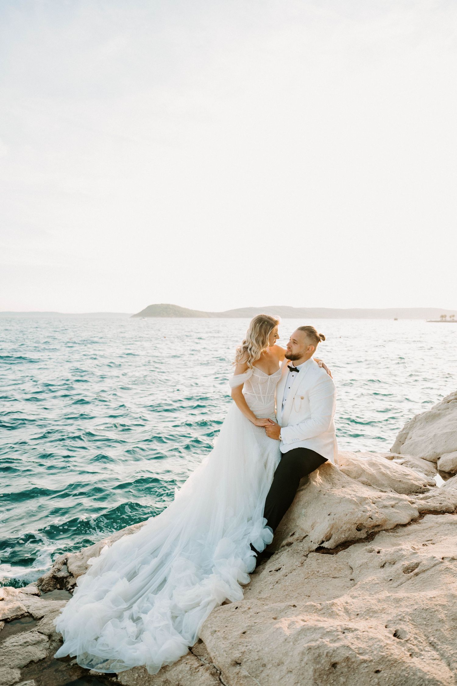 A couple in wedding attire embraces on coastal rocks overlooking a serene ocean view at sunset.