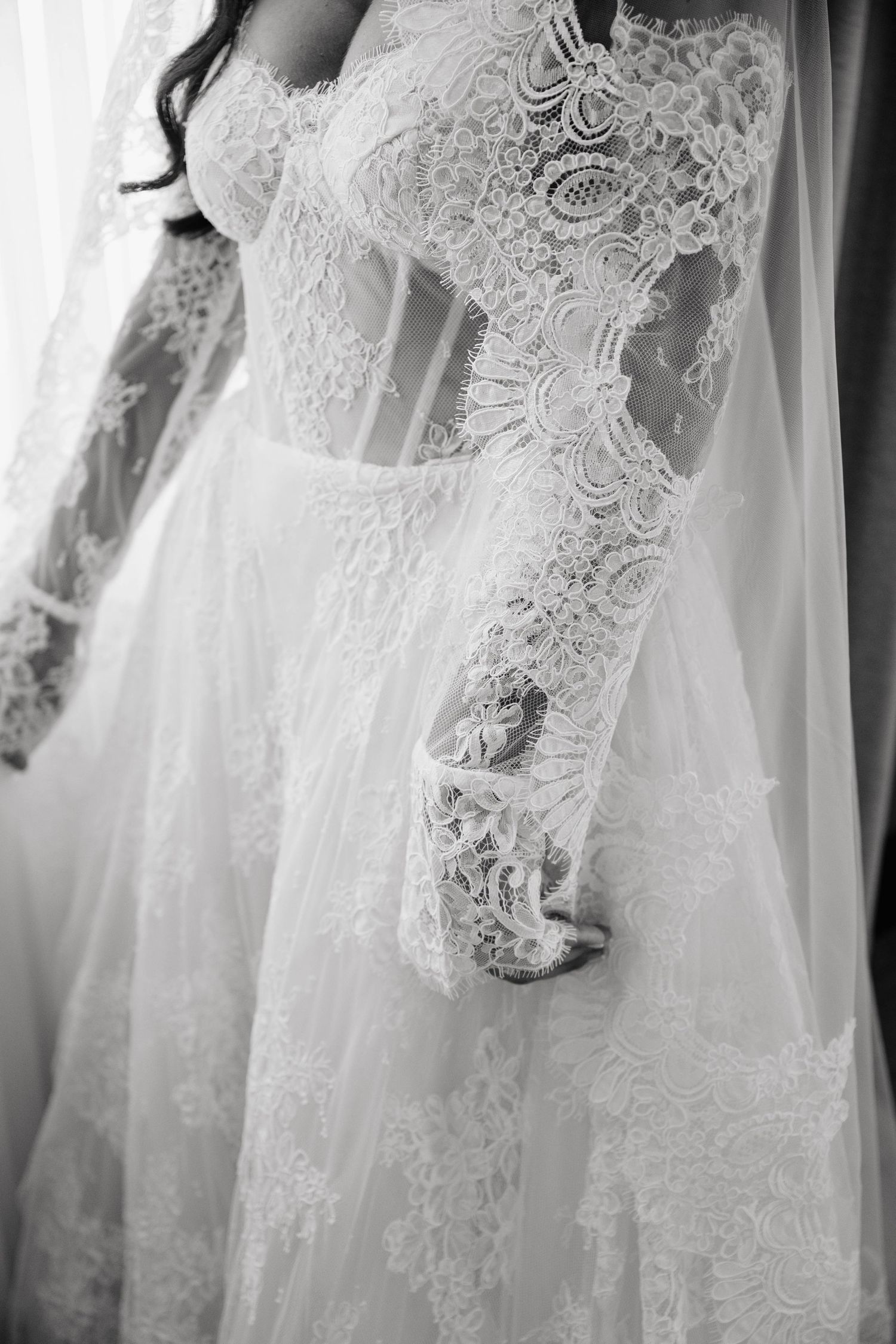 Black and white close-up of a bride in an elegant long-sleeve lace wedding gown with intricate floral detailing.