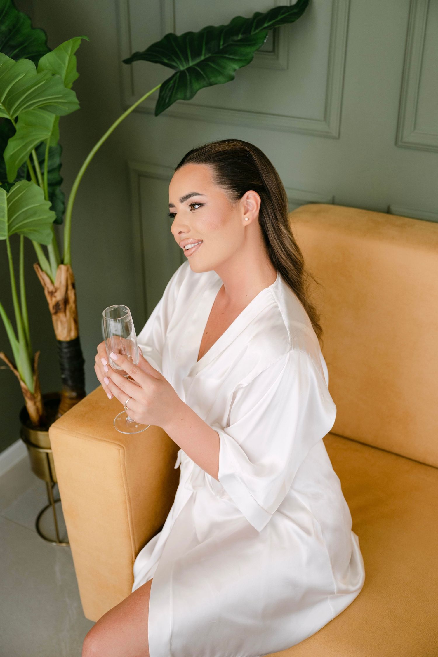 Woman in white satin robe holding champagne flute, seated on gold velvet sofa near tropical plant in elegant green room.