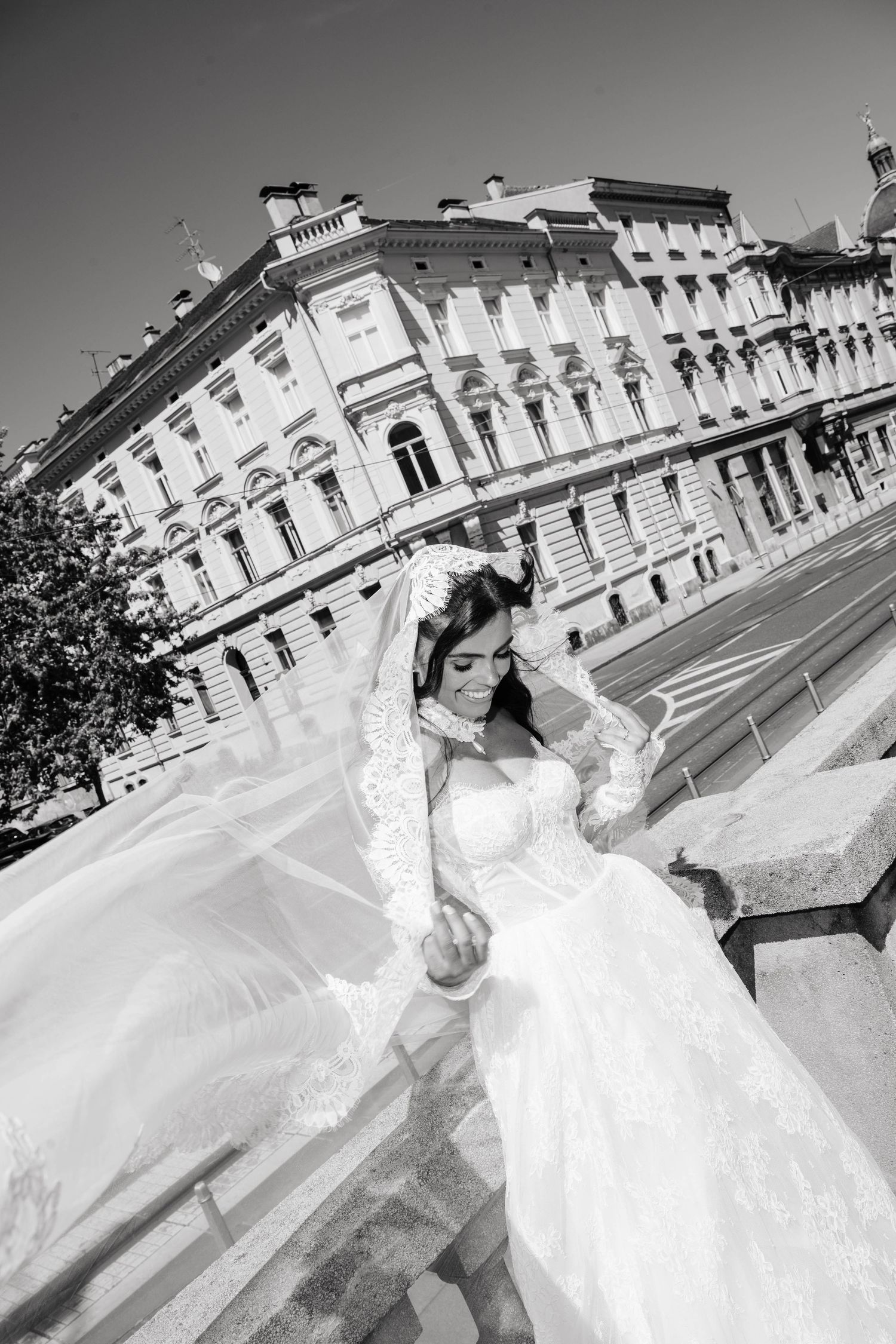 Black and white photo of a bride in a flowing lace wedding dress and veil walking near a grand European building.