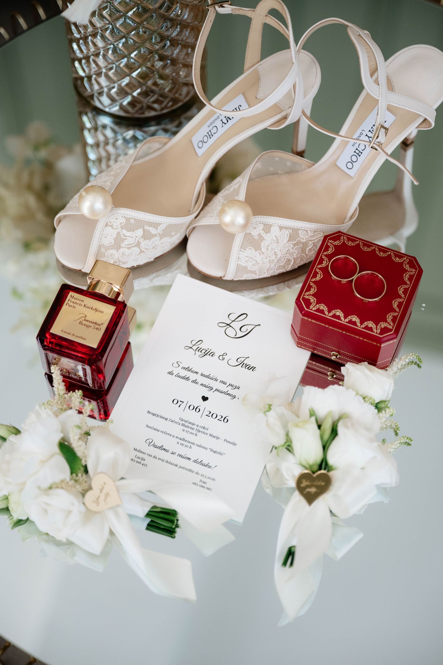 Elegant wedding flat lay with lace bridal heels, gold rings in red box, perfume, white flowers, and invitation dated 07.06.2020.
