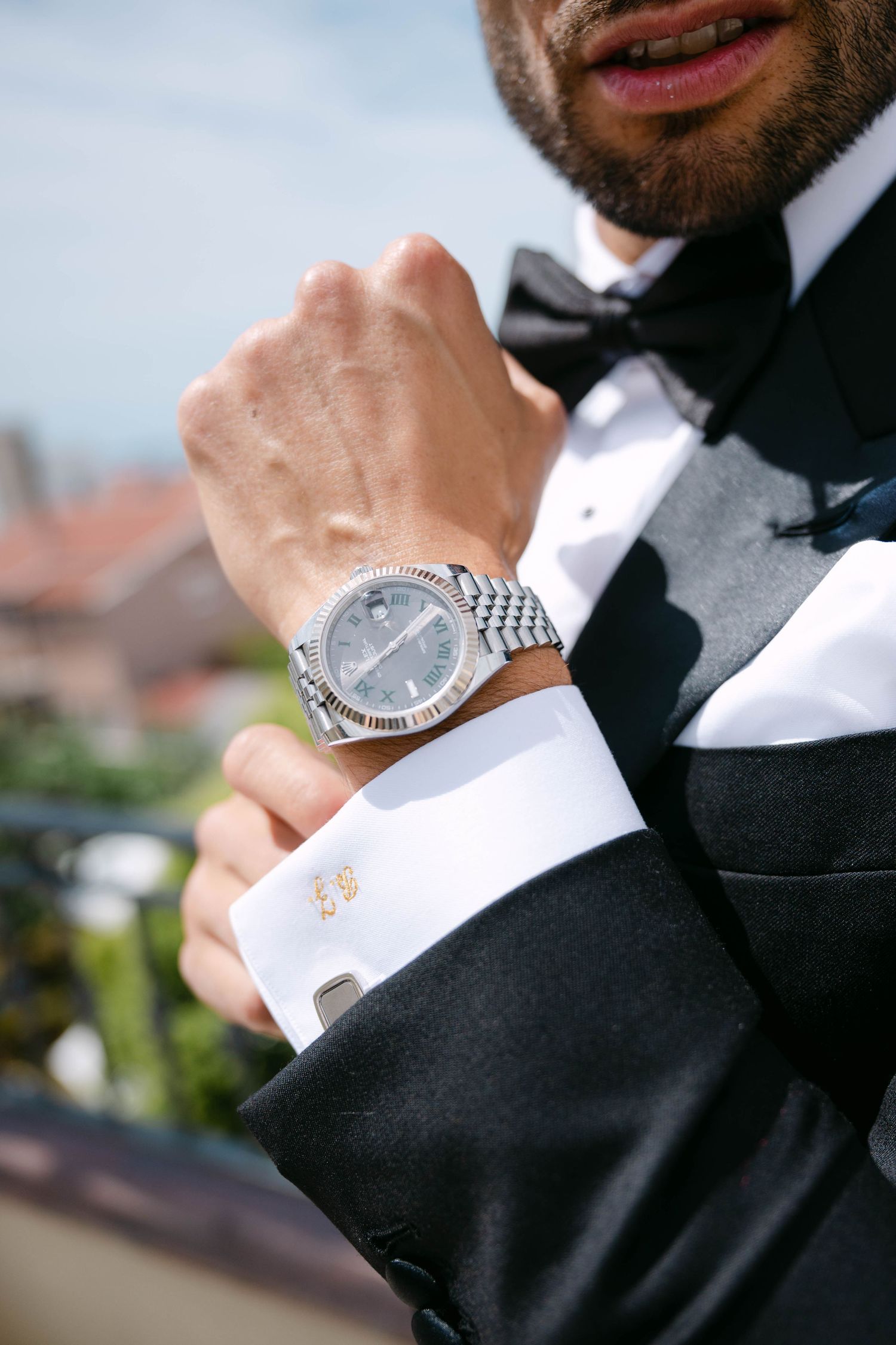 Groom in black tuxedo adjusting his luxury silver Rolex watch with white French cuff shirt and bow tie.