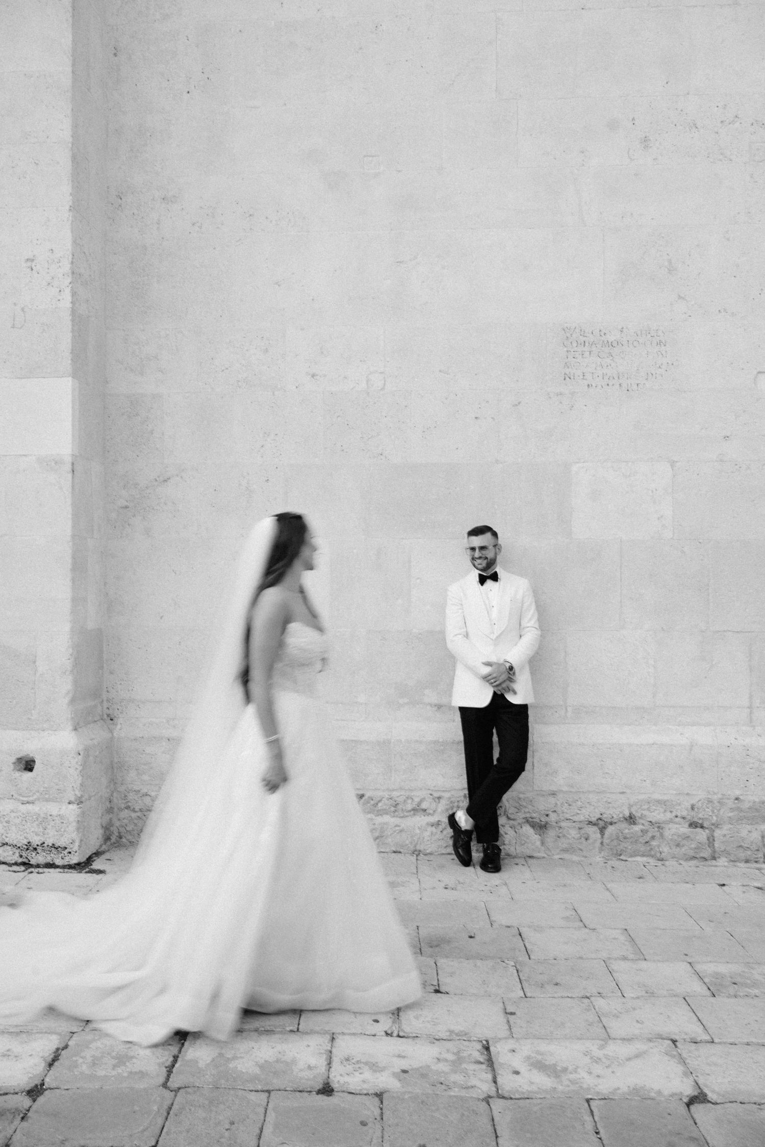 Black and white wedding photo of bride in flowing gown approaching groom in white tuxedo leaning against stone wall.