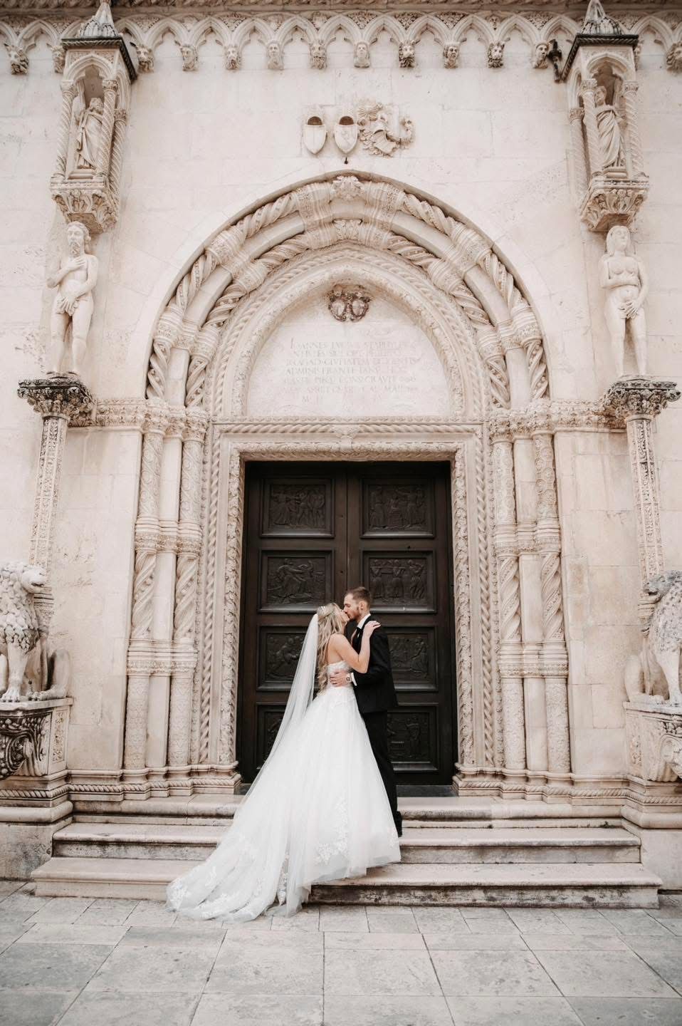 Wedding portrait in front of ornate stone church doorway with detailed Gothic architectural elements.