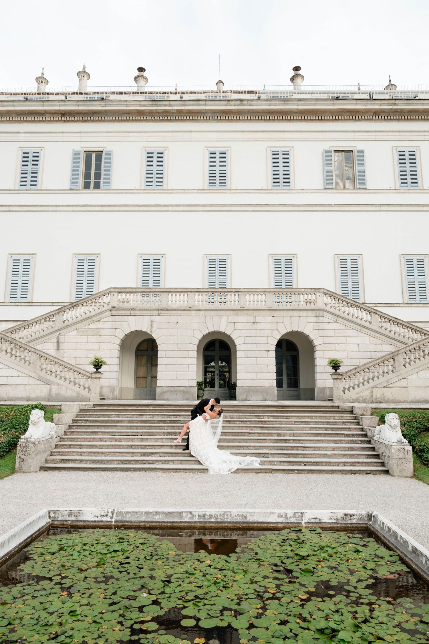 Bride and groom embrace on grand marble steps of elegant white Italian villa with lily pond in foreground.