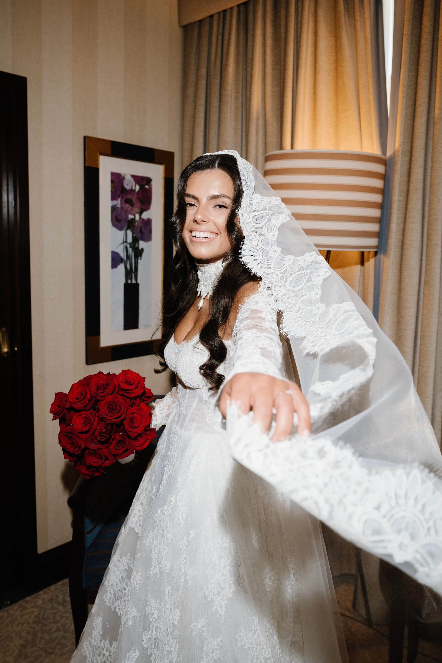 Smiling bride in lace wedding gown and veil holds red rose bouquet in elegant room.