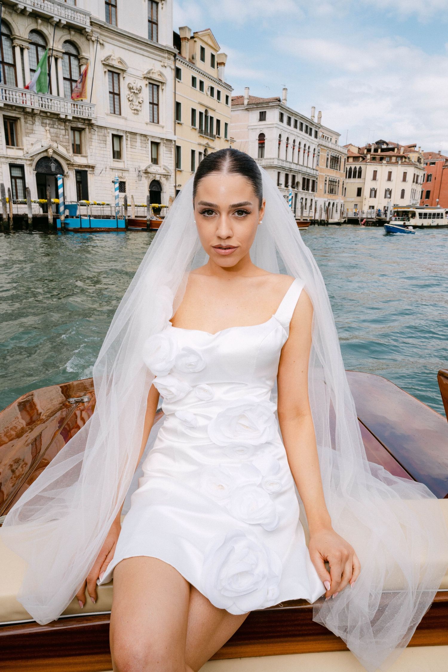 Bride in short white dress and flowing veil sits on wooden boat in Venice canal with historic buildings in background.