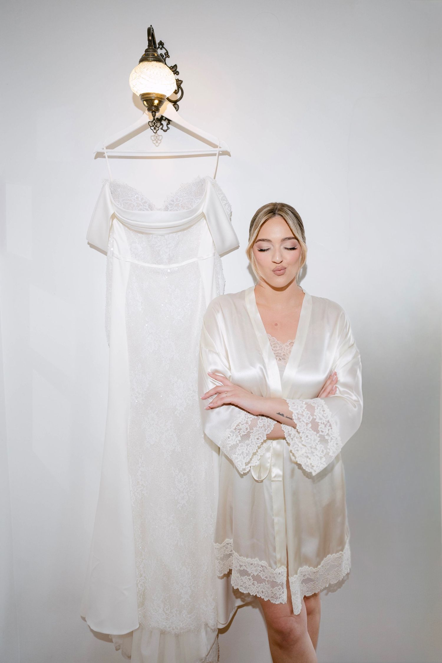 Bride in satin robe standing beside elegant white lace wedding dress hanging on wall sconce.