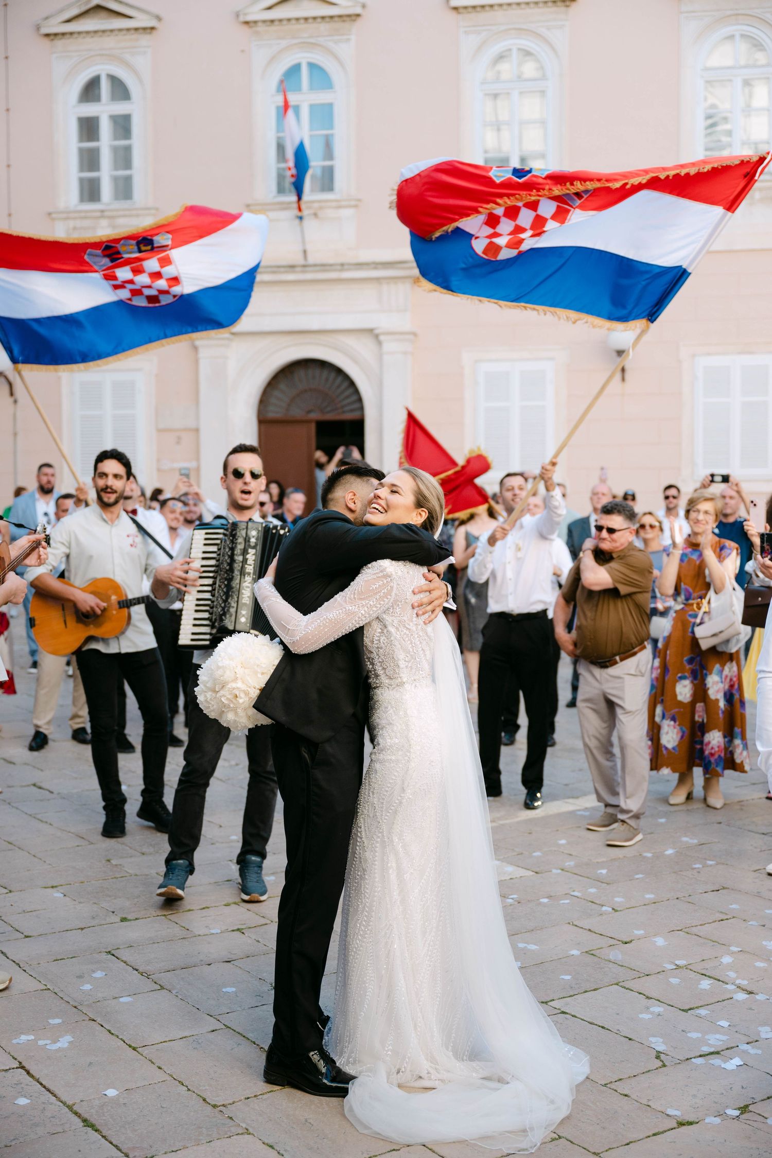 Bride and groom embrace at Croatian wedding celebration with flags and accordion player in sunny town square.