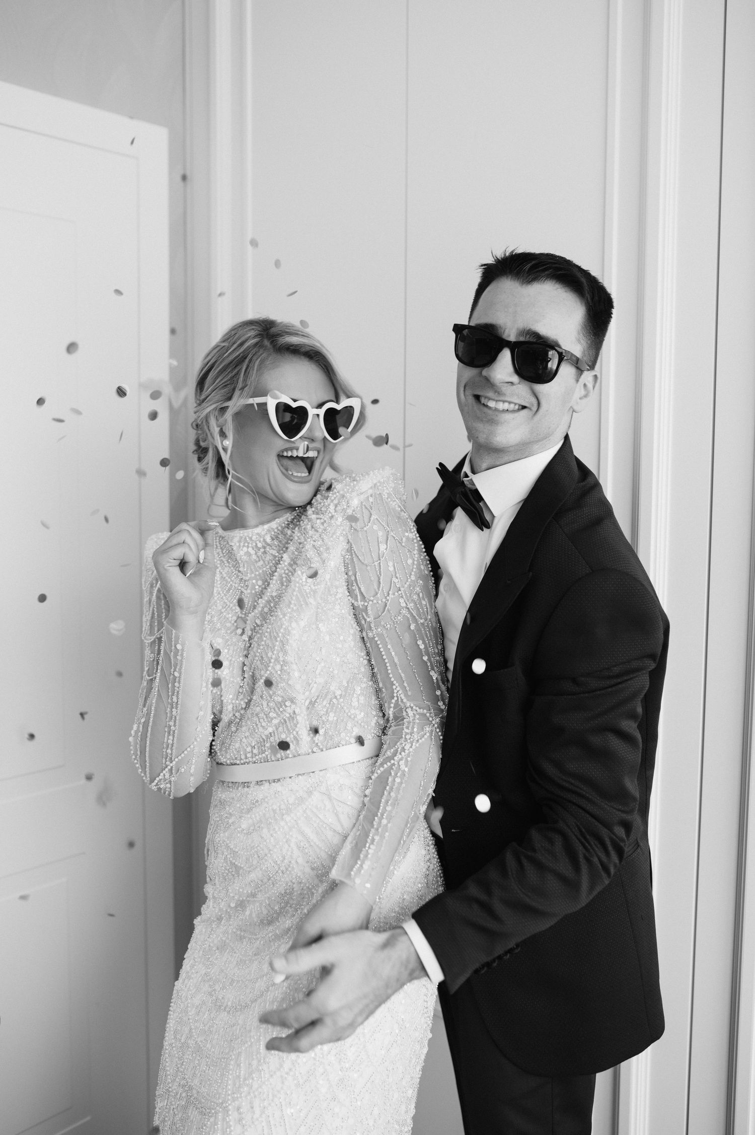 Stylish couple in wedding attire wearing sunglasses pose playfully together.