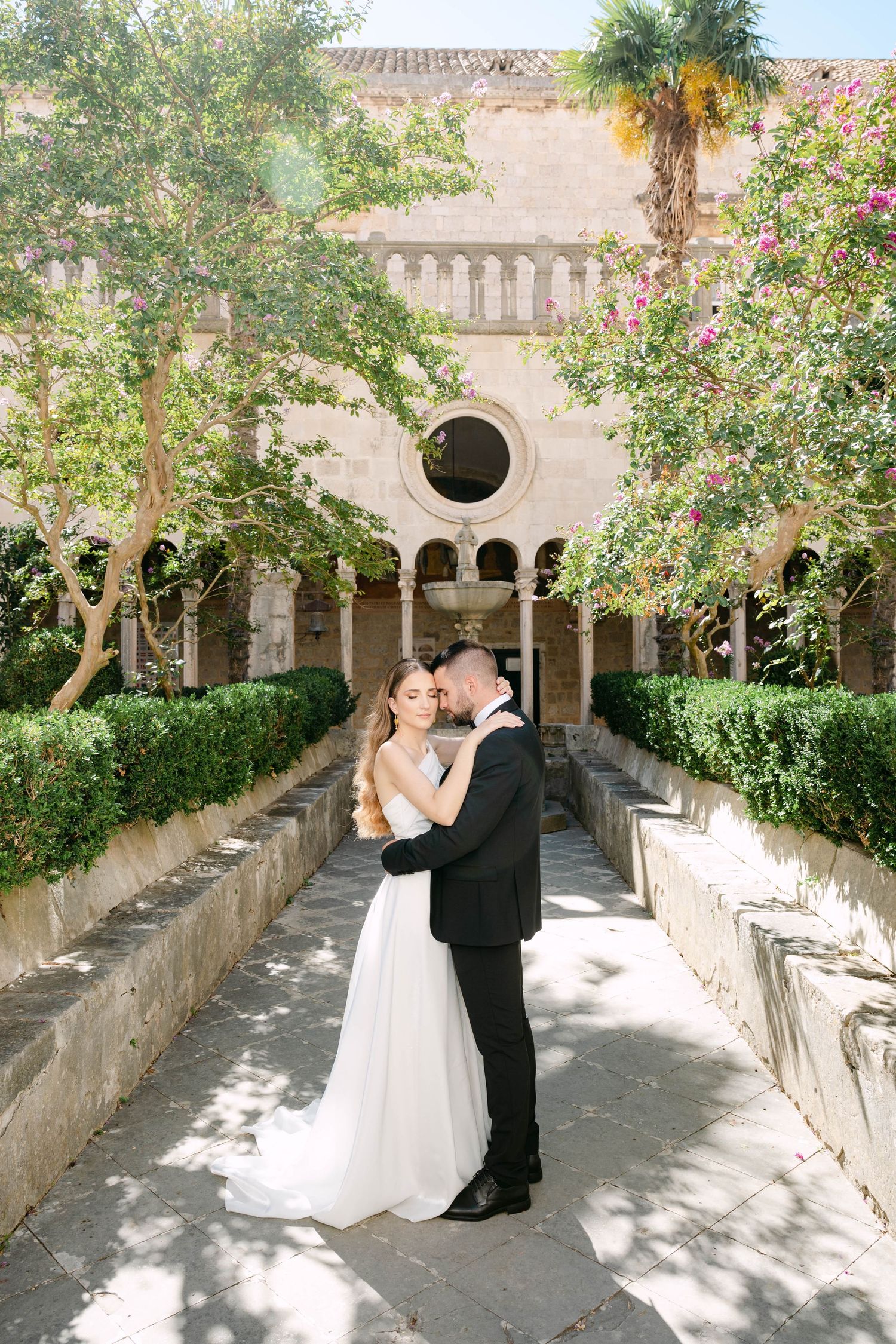 A romantic couple shares tender moments in a Mediterranean courtyard with stone arches and blooming flowers.
