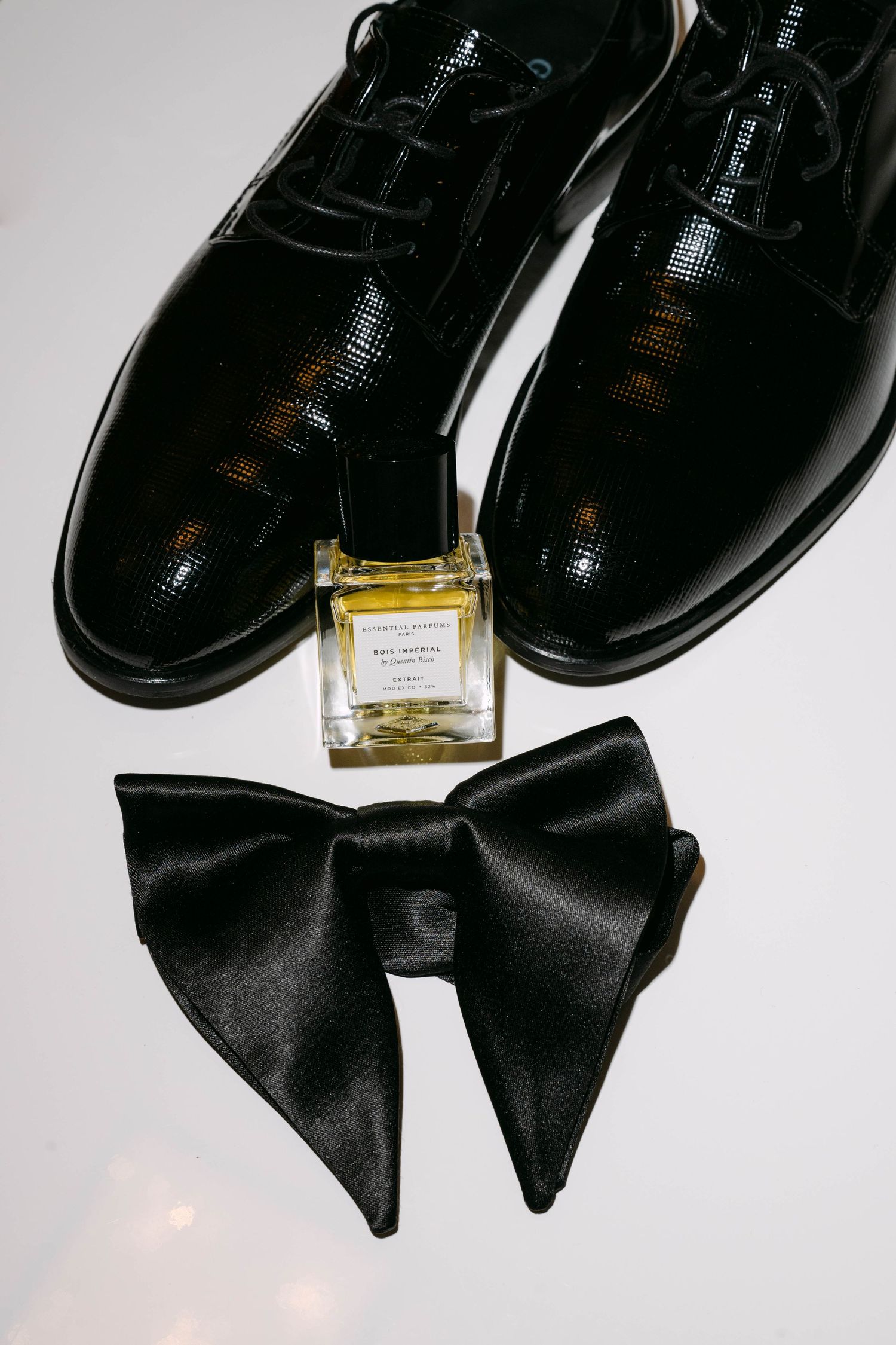 Black patent leather dress shoes, bow tie, and cologne arranged on white surface for a wedding.