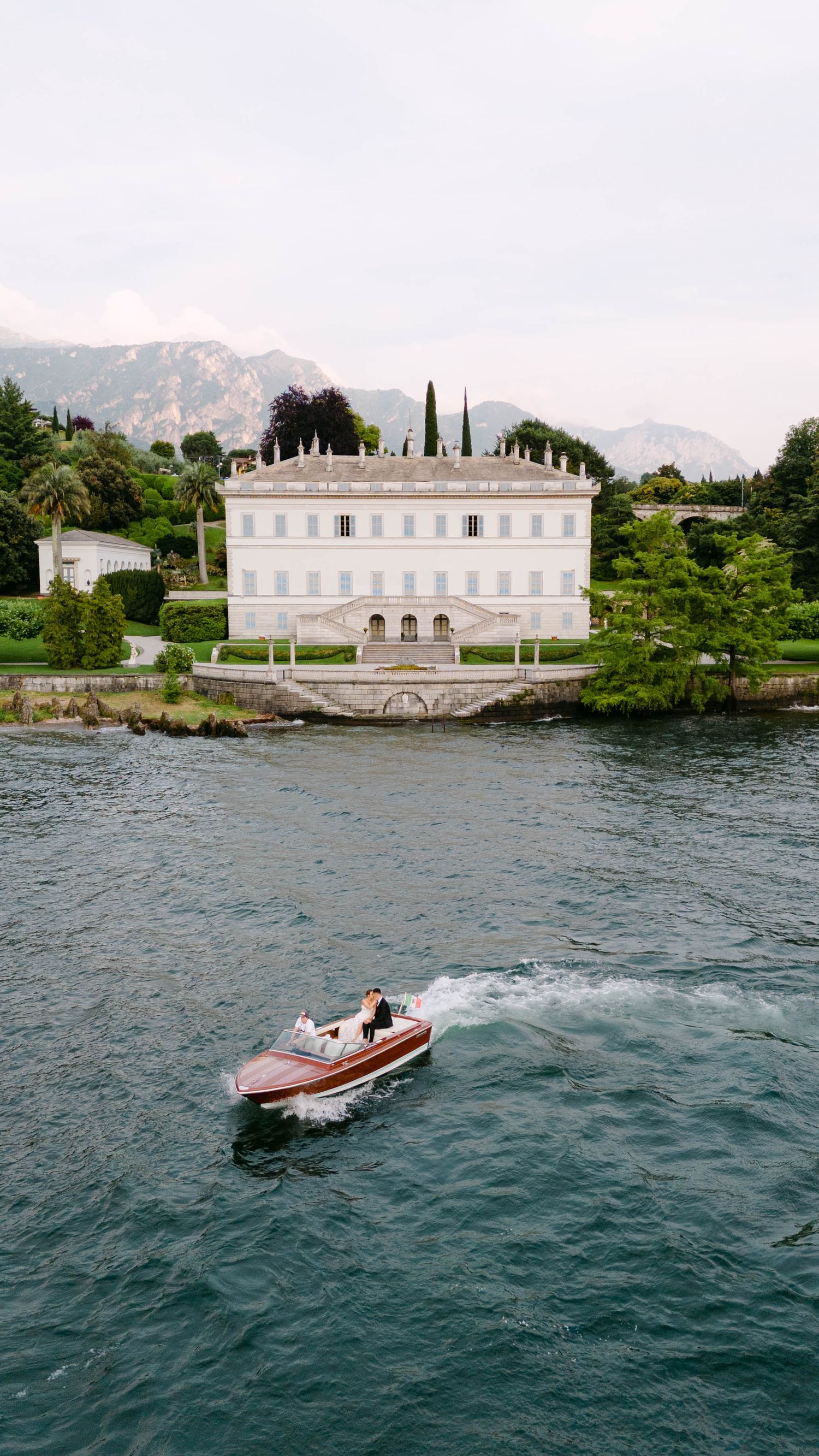 Couple riding a classic wooden speedboat on Lake Como with an elegant Italian villa and mountain backdrop.