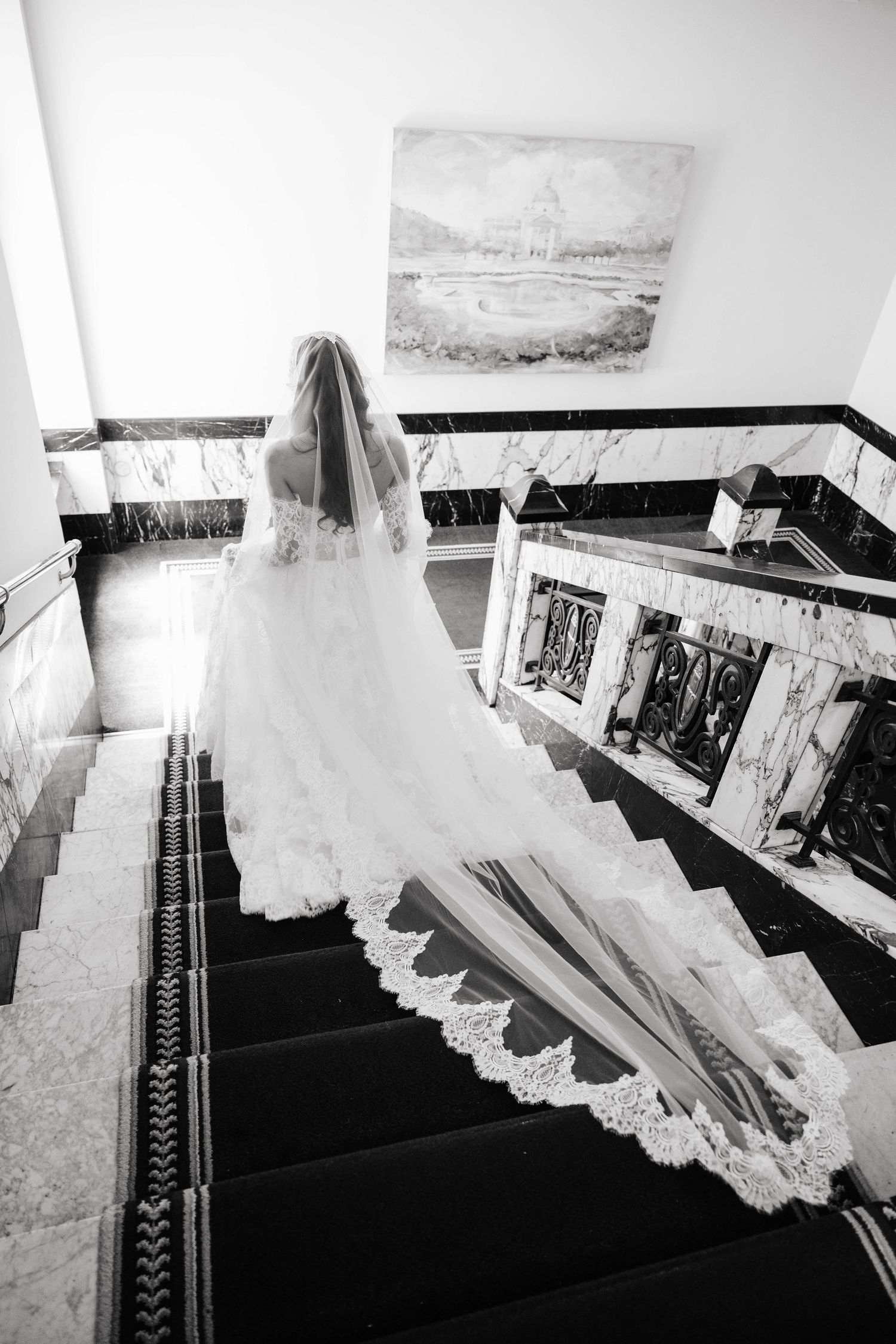 Black and white photo of a bride ascending marble stairs, her lace wedding gown and cathedral veil trailing elegantly behind her.