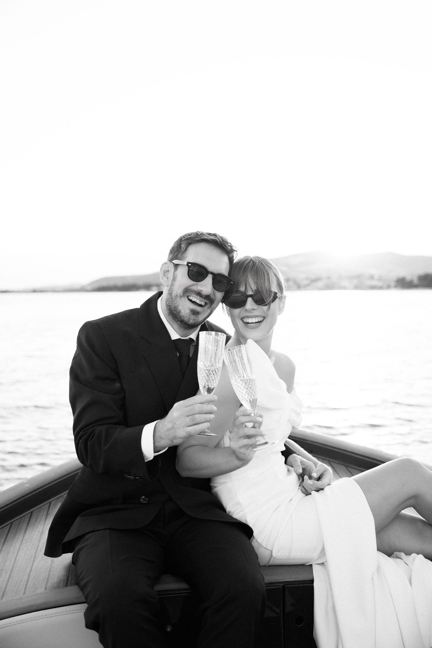 A happy couple toasts with champagne glasses while sitting on a boat in black and white photography.