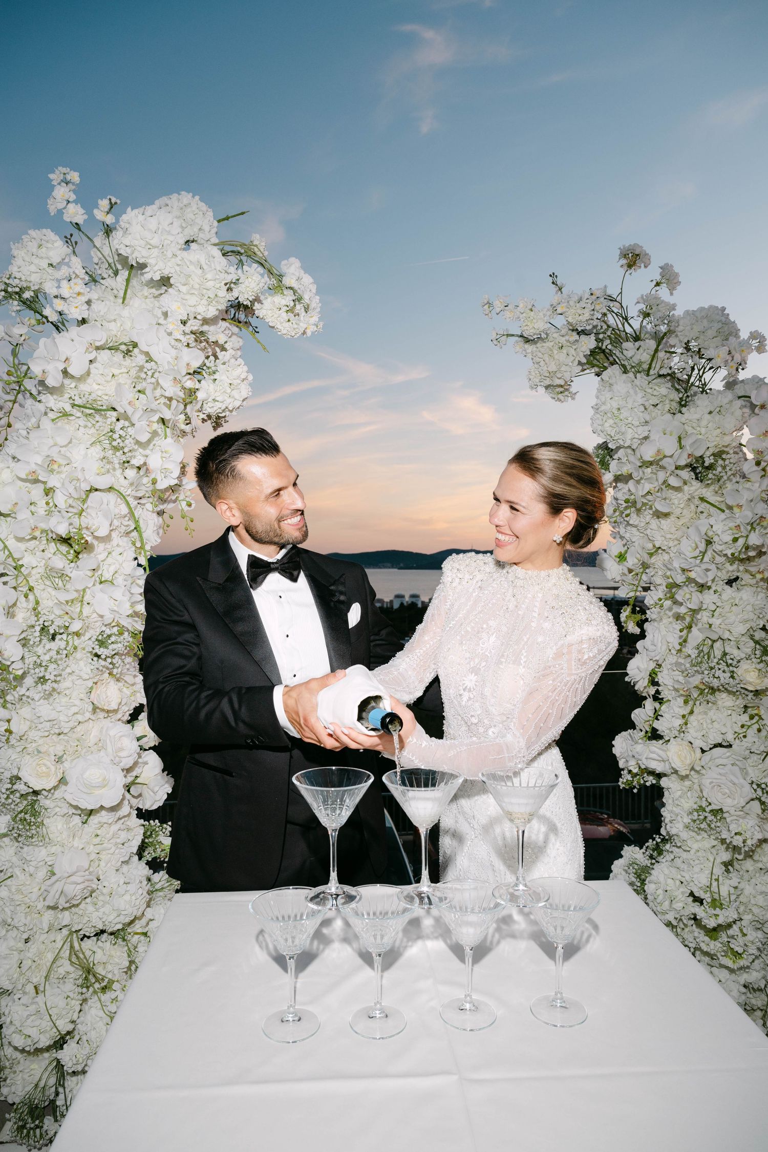 Bride and groom pouring champagne tower at elegant wedding reception surrounded by white floral arrangements at sunset.