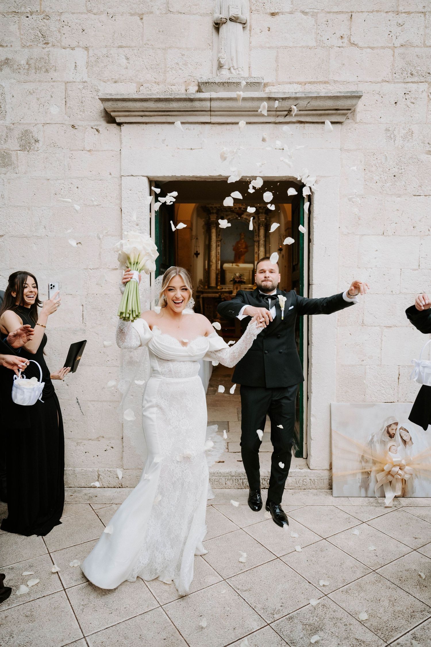 Bride and groom exit stone church amid flower petal confetti toss, bridesmaids in black dresses celebrating joyful wedding ceremony.