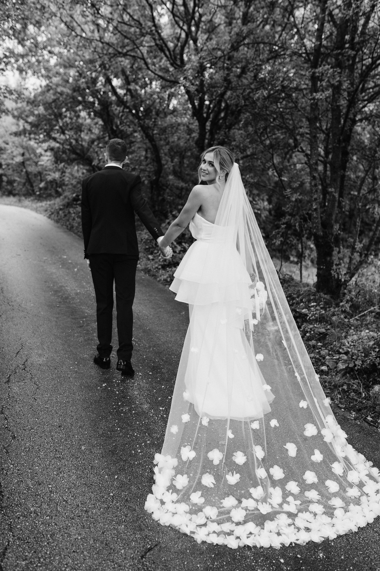 A bride in a flowing white dress with floral lace veil walks down a wooded path behind her groom in black suit.