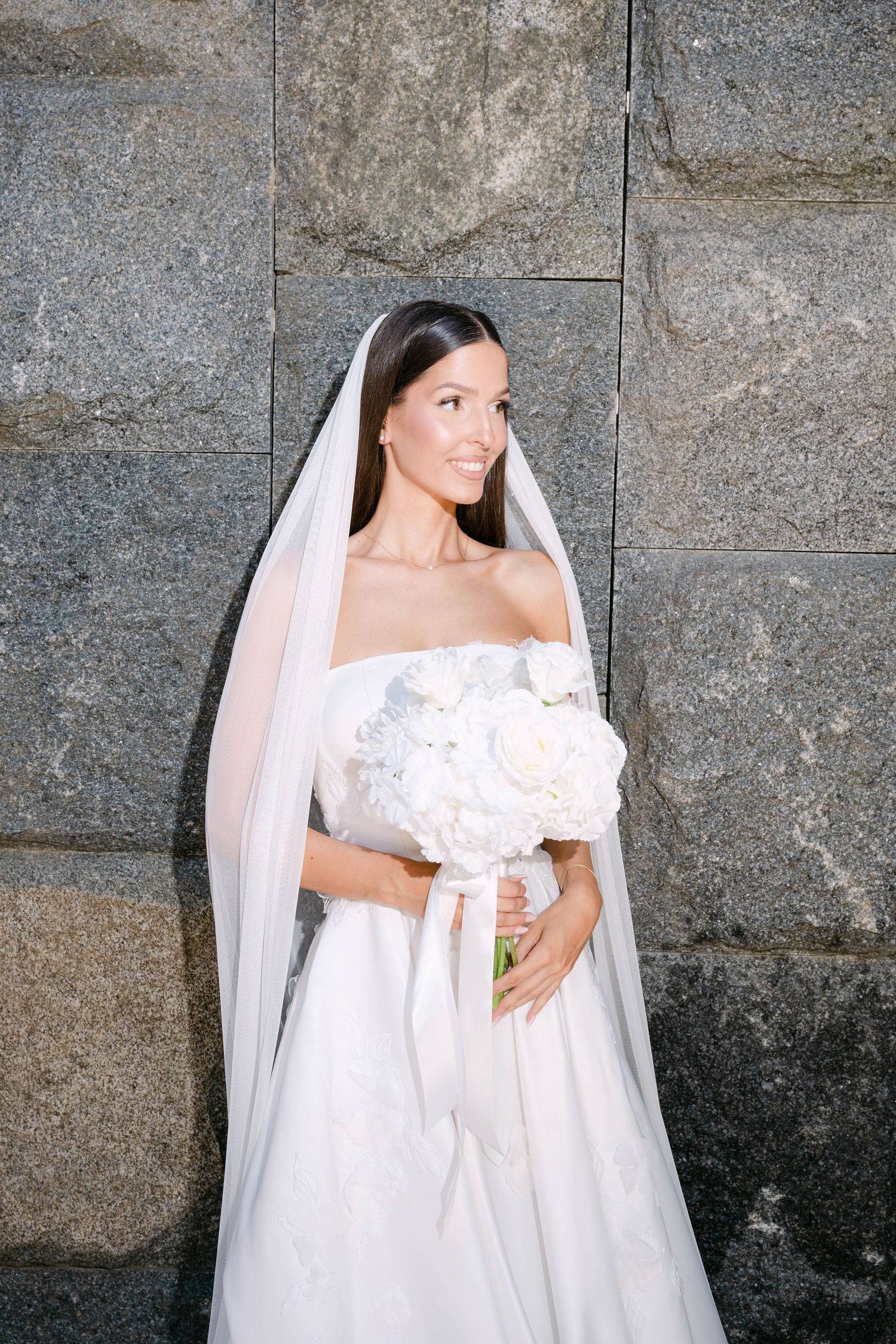 A bride in an elegant off-shoulder white wedding gown and veil poses against a textured stone wall.