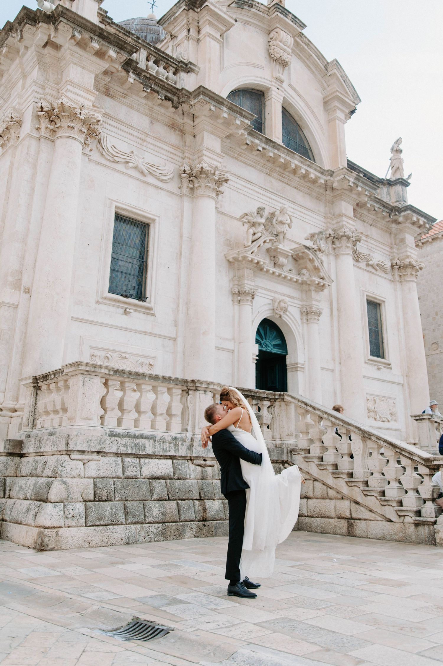 A couple embraces on the grand stone steps of an ornate white Baroque church with decorative architectural details.