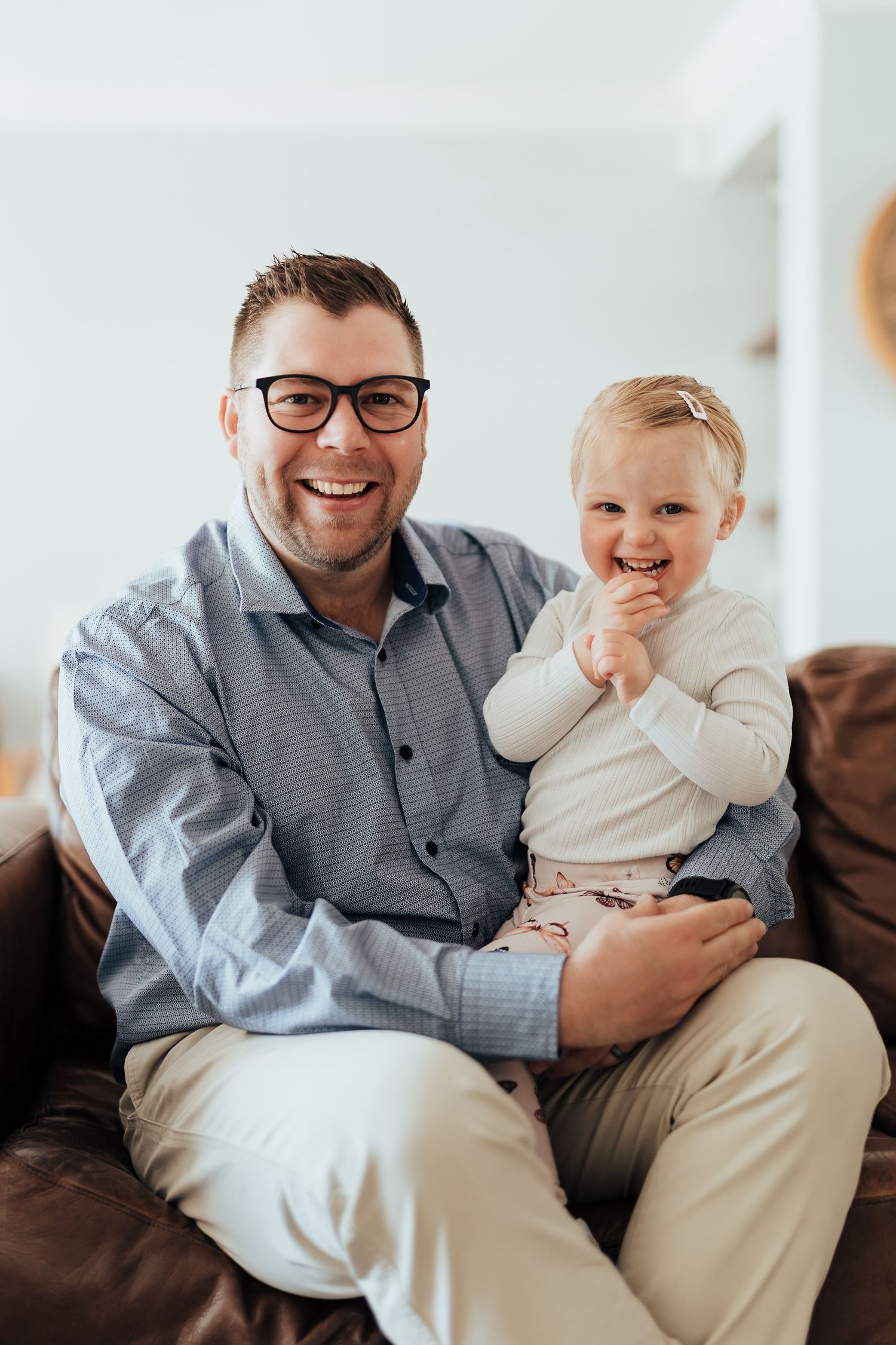 A person in a gray shirt sits on a couch, smiling and interacting with a small child wearing white clothing.