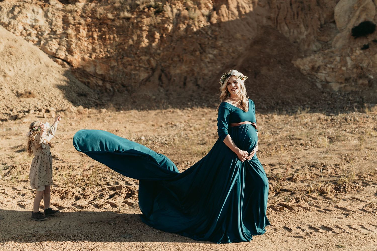 Elegant teal flowing maternity gown billows in the wind against a desert canyon backdrop.