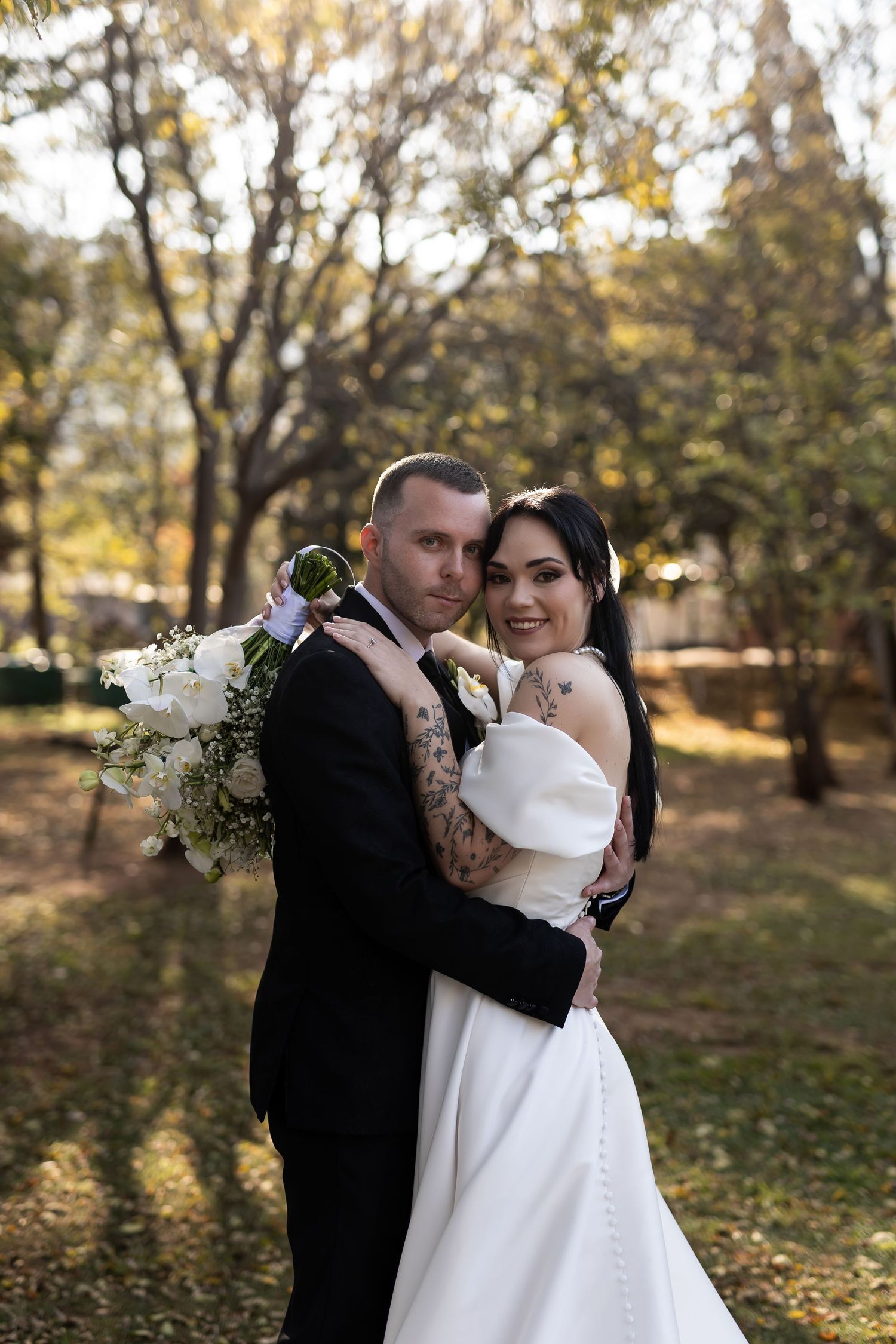 A wedding couple embraces joyfully in an autumn forest setting with golden leaves and sunlight filtering through trees.