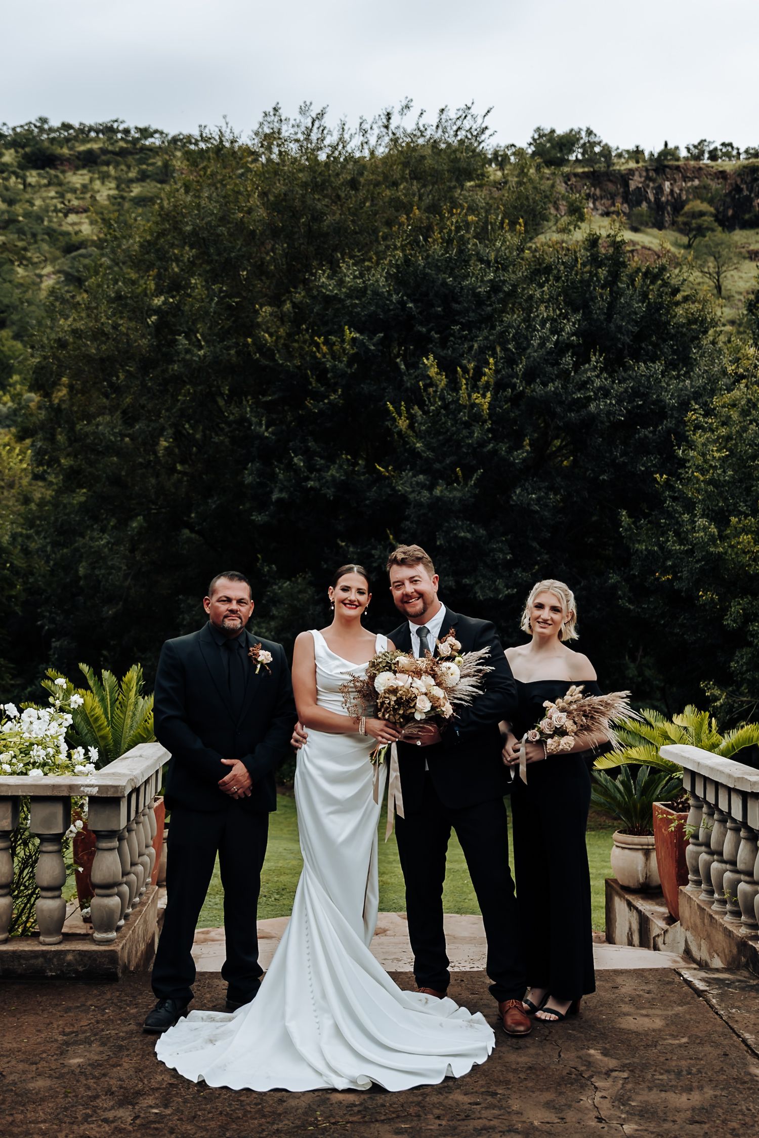 Wedding party poses together on a rustic stone balcony surrounded by lush greenery and trees.