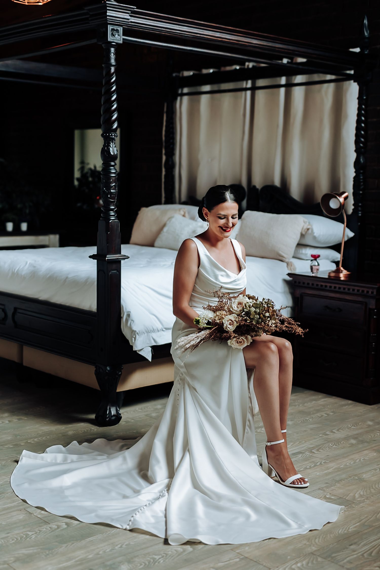 A white wedding dress flows elegantly across a dark wooden floor in a bedroom with four-poster bed.
