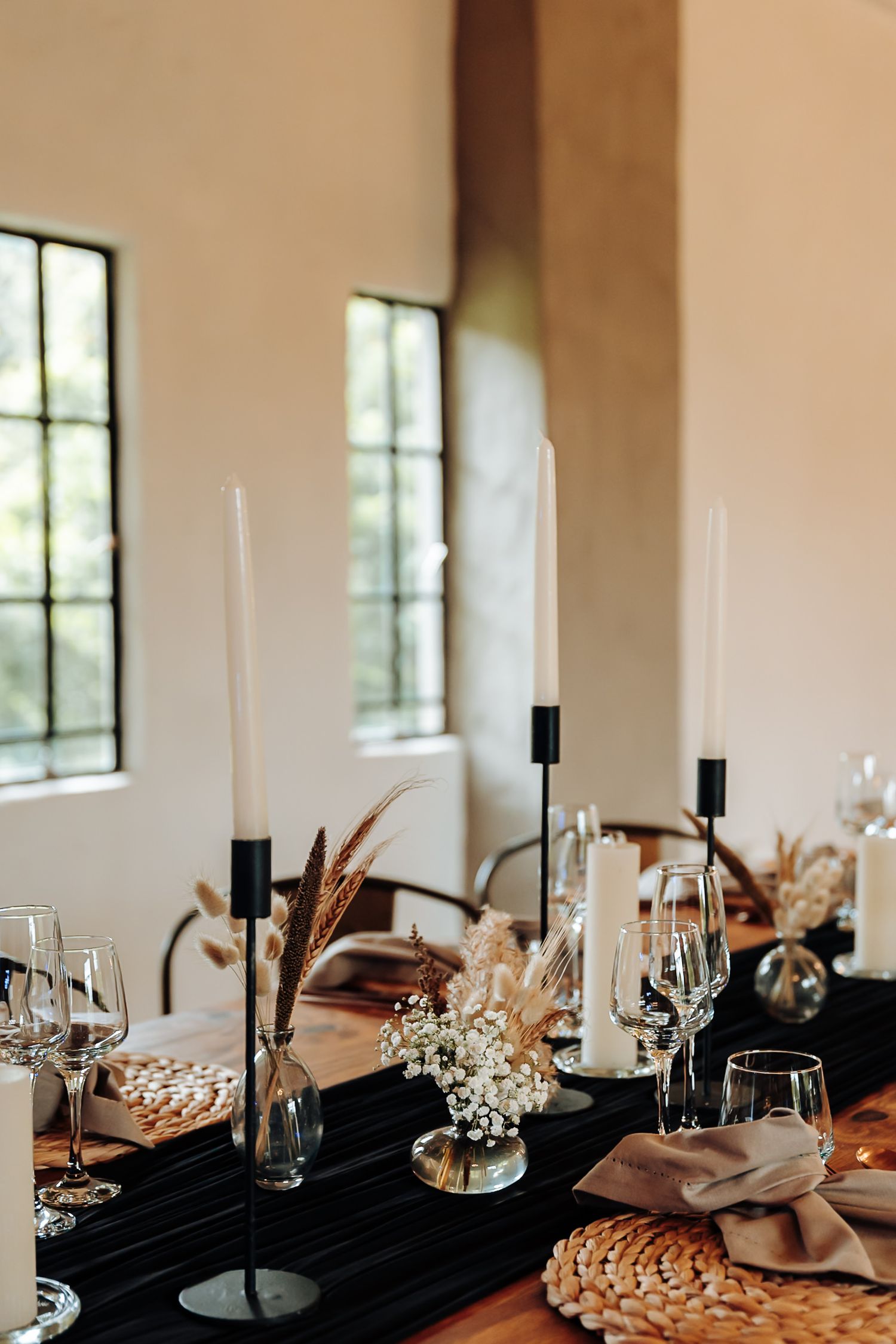 Elegant wedding table setting with black candles, dried flowers, and crystal glassware in a bright modern venue.
