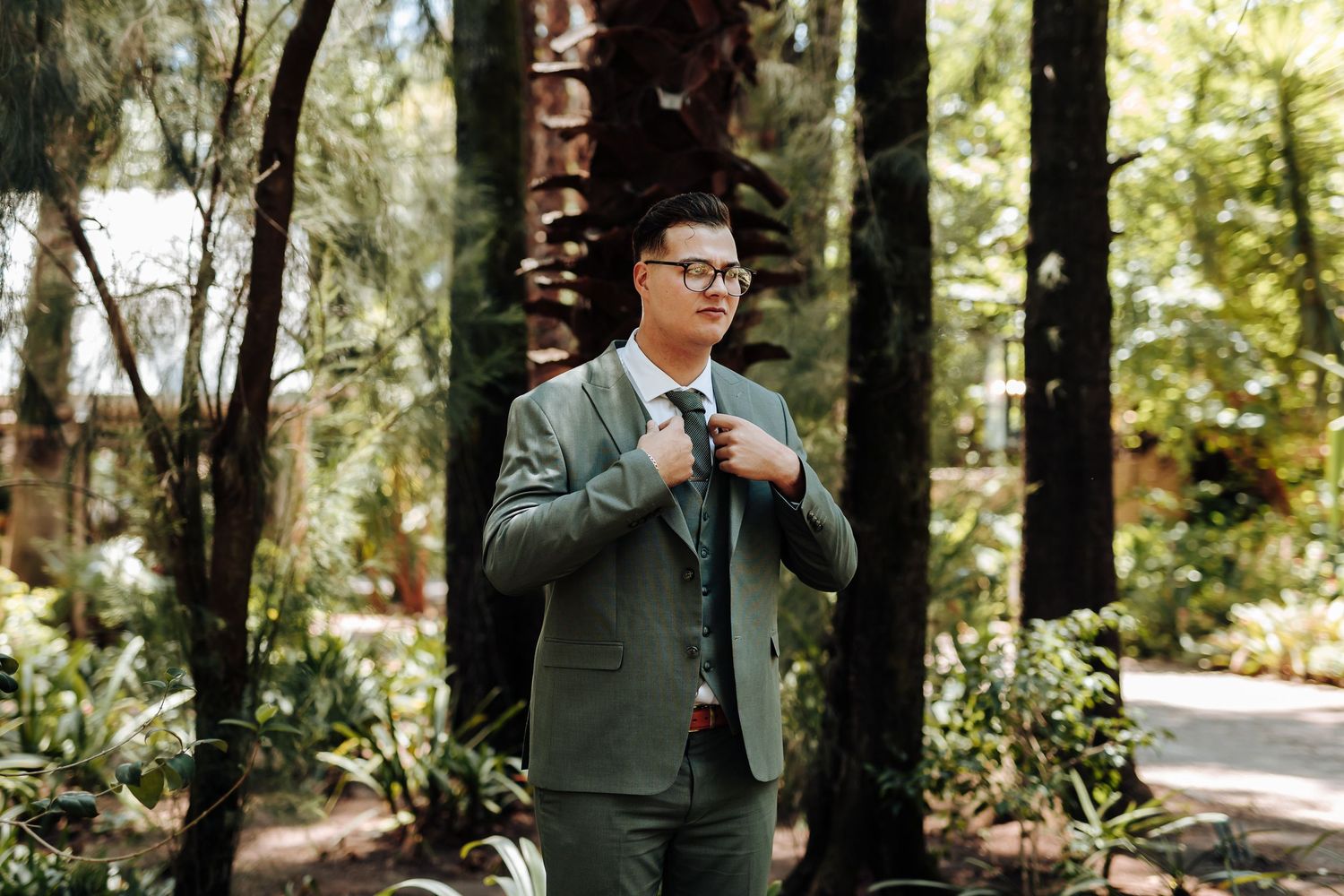 A person in a green jacket poses thoughtfully among tall trees in a serene forest setting.