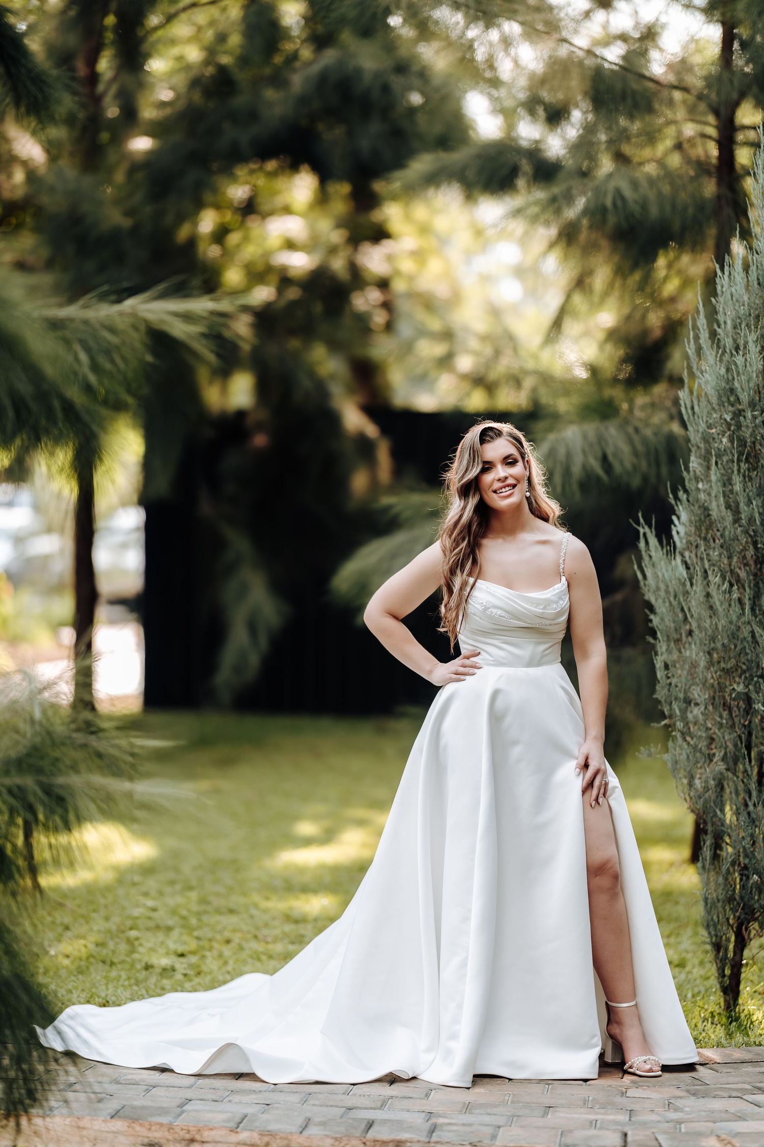 Elegant bride in a flowing white gown poses gracefully in a sunlit garden setting with tall evergreen trees.