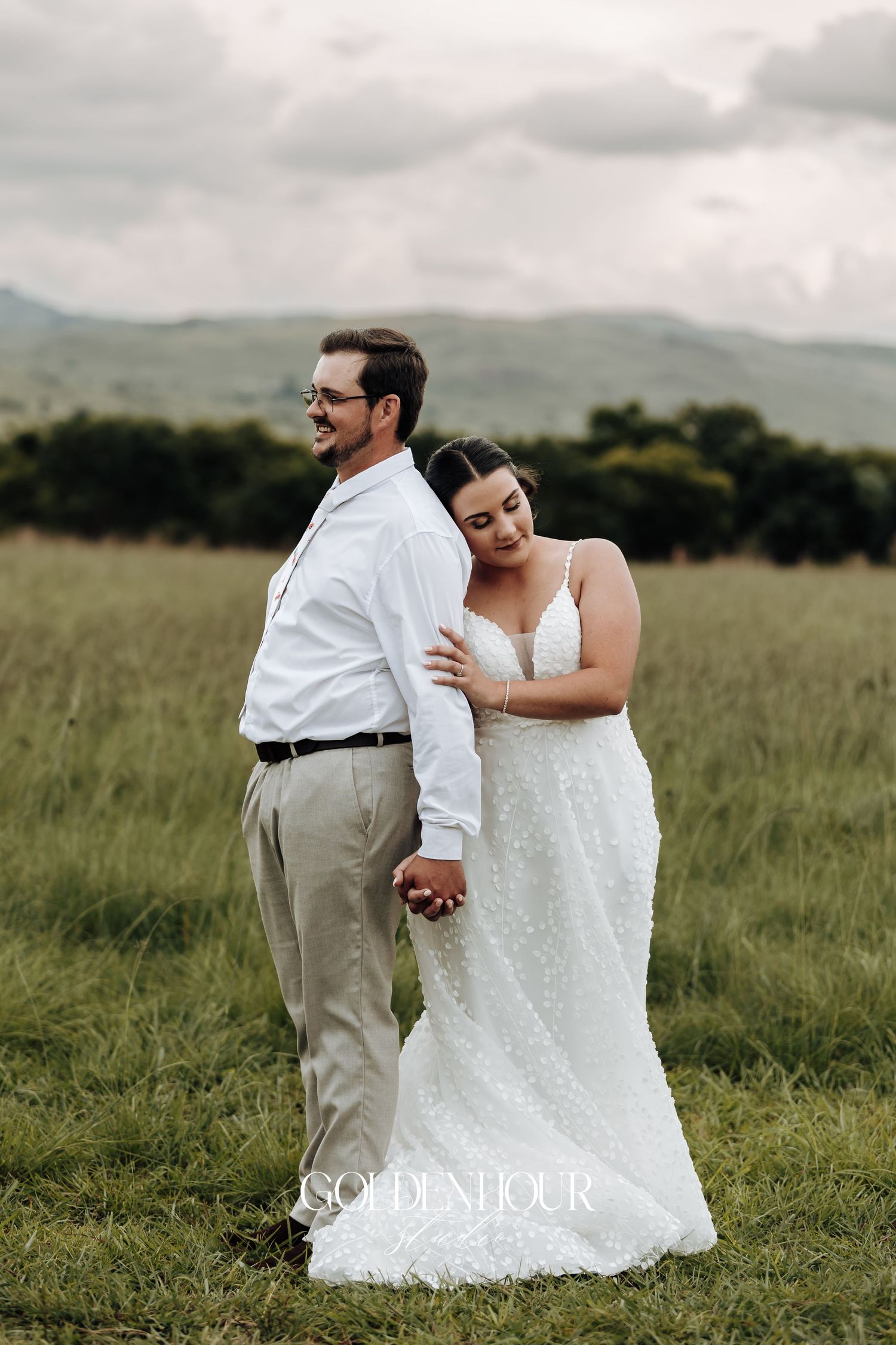 A couple in wedding attire shares an intimate moment together in a scenic grassy field with mountains in the background.