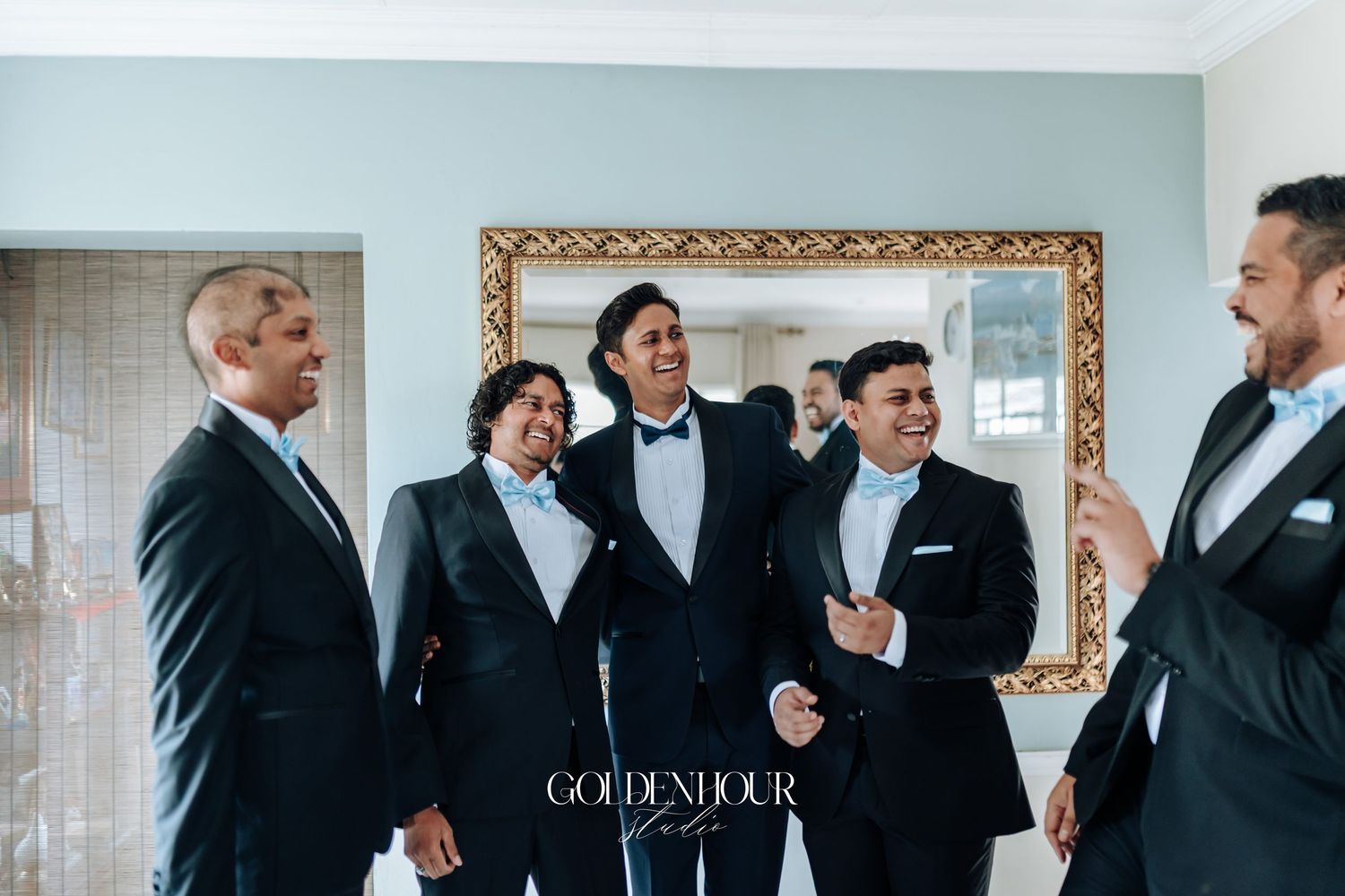 Wedding party in formal black suits and dresses gather for candid moments in elegant room with gold mirror.