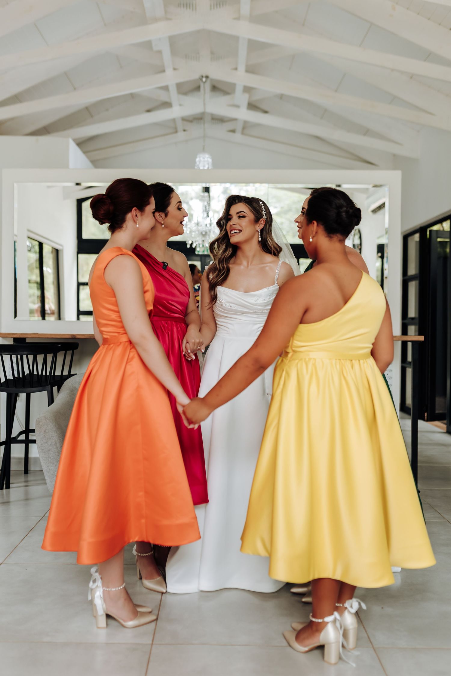 Bridesmaids in colorful orange, red, and yellow dresses gather together in a bright indoor venue.