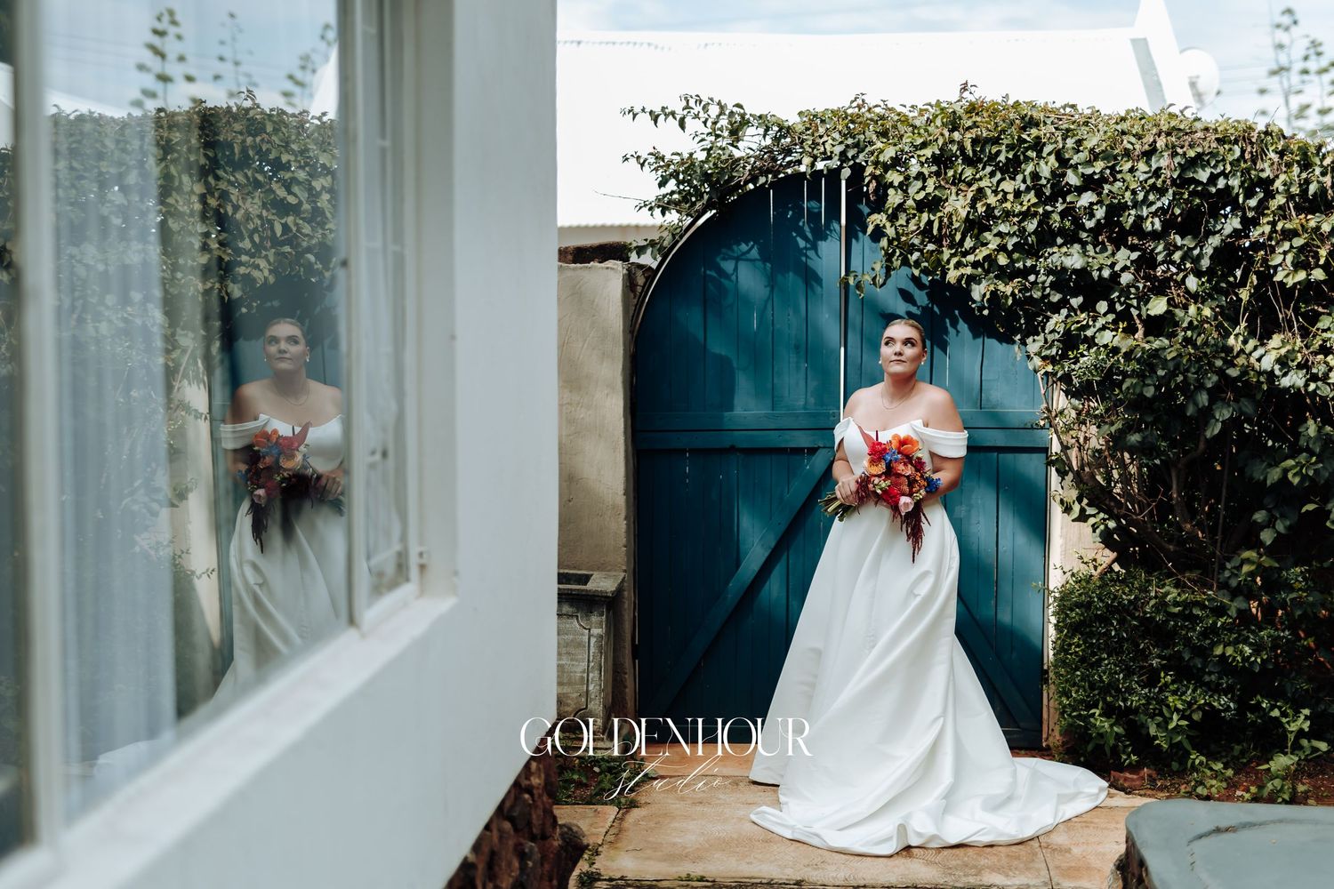 A bride in a white wedding dress poses in front of a blue door with greenery and flowers in the background.