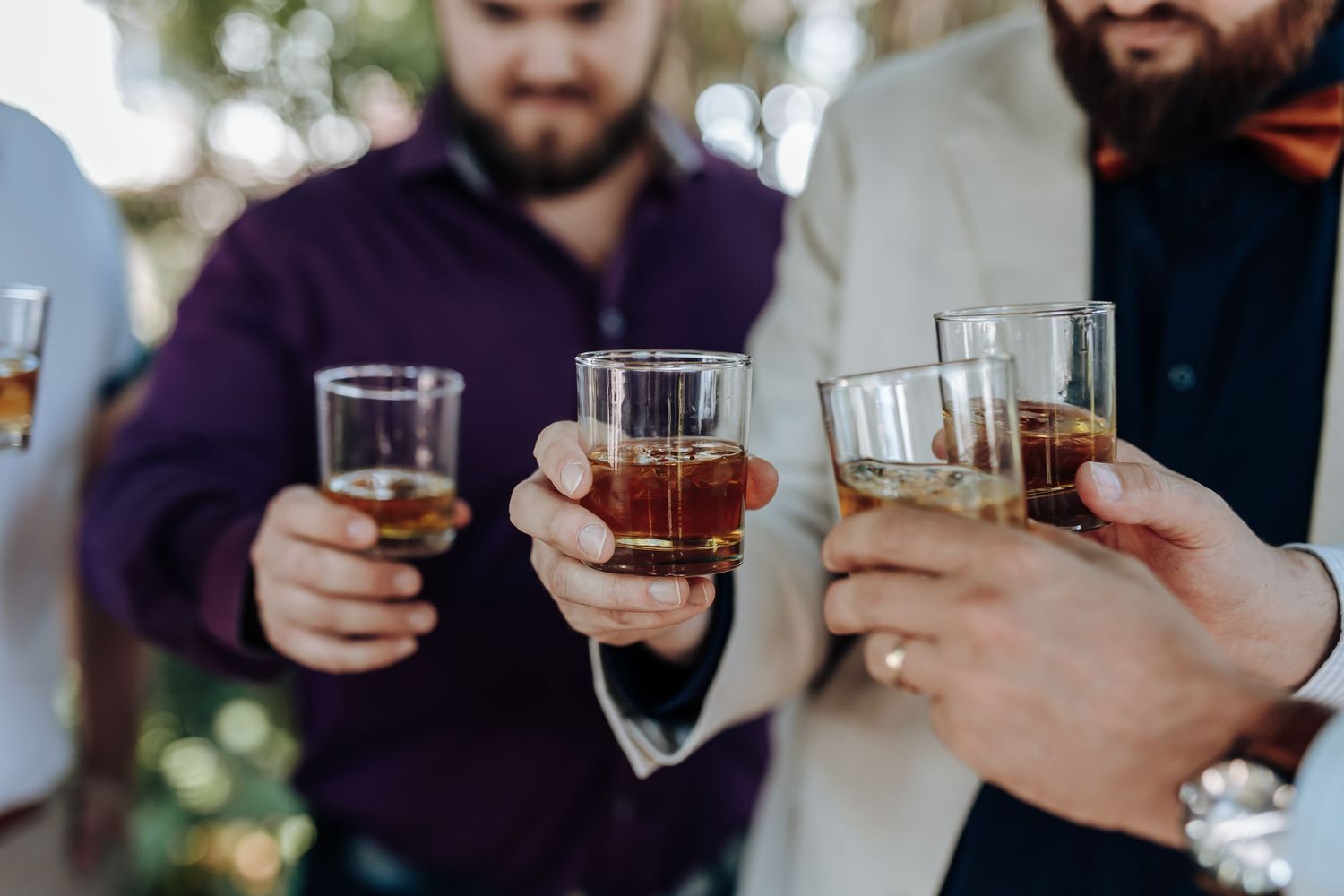 People clink glasses together in a toast at a social gathering while holding drinks and phones.