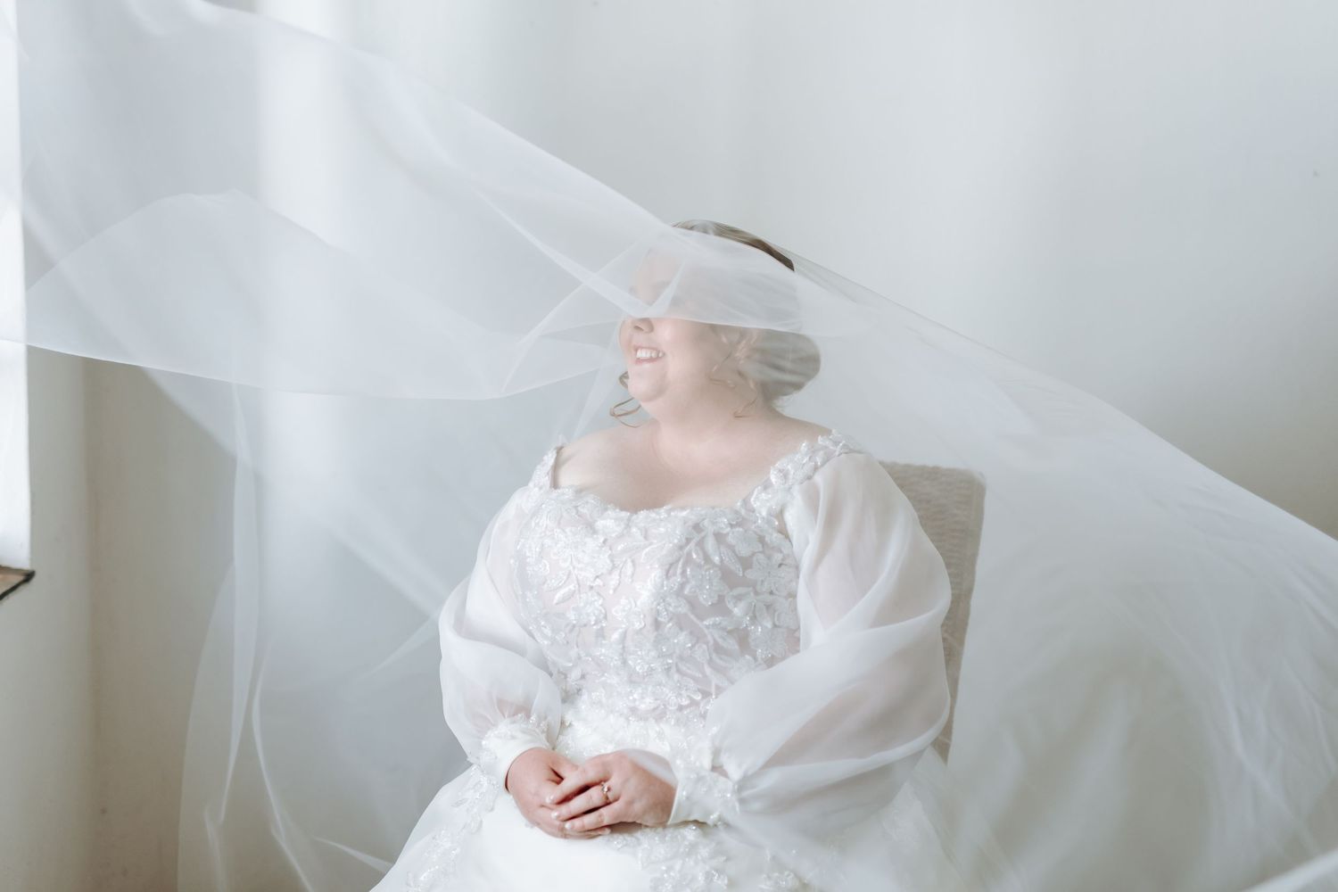 A bride in a white lace wedding dress poses with flowing veil material creating an ethereal bridal portrait.