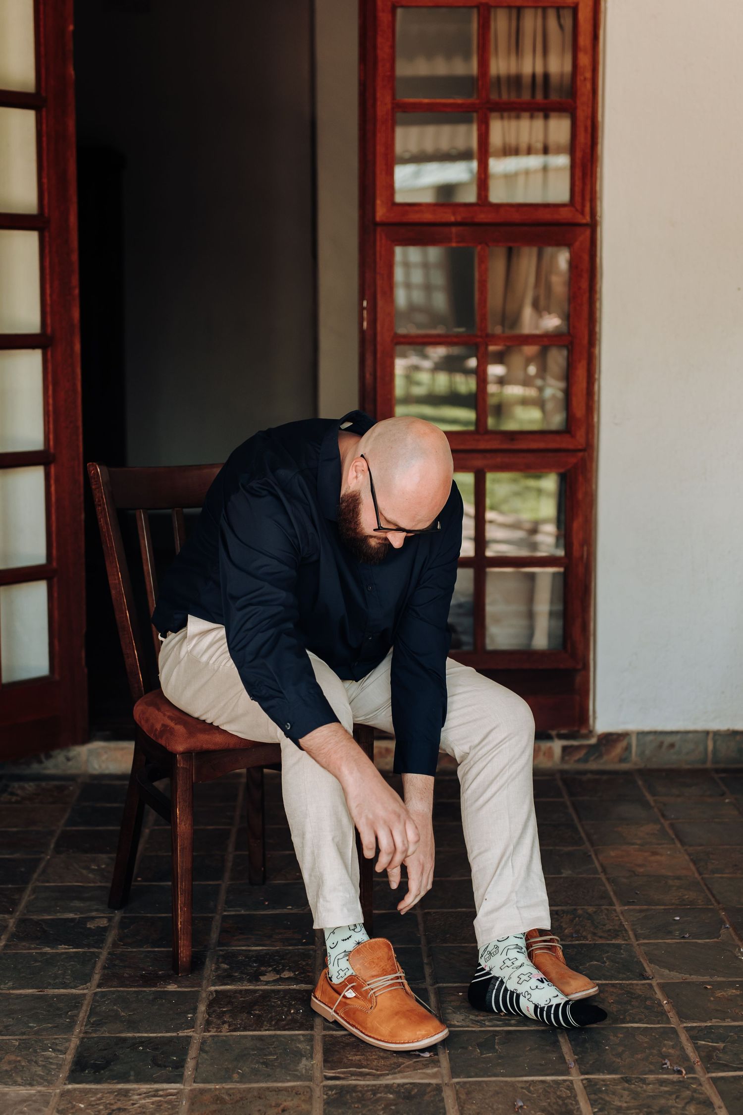 A person in a navy blazer and light pants sits on a wooden chair while tying brown leather shoes.