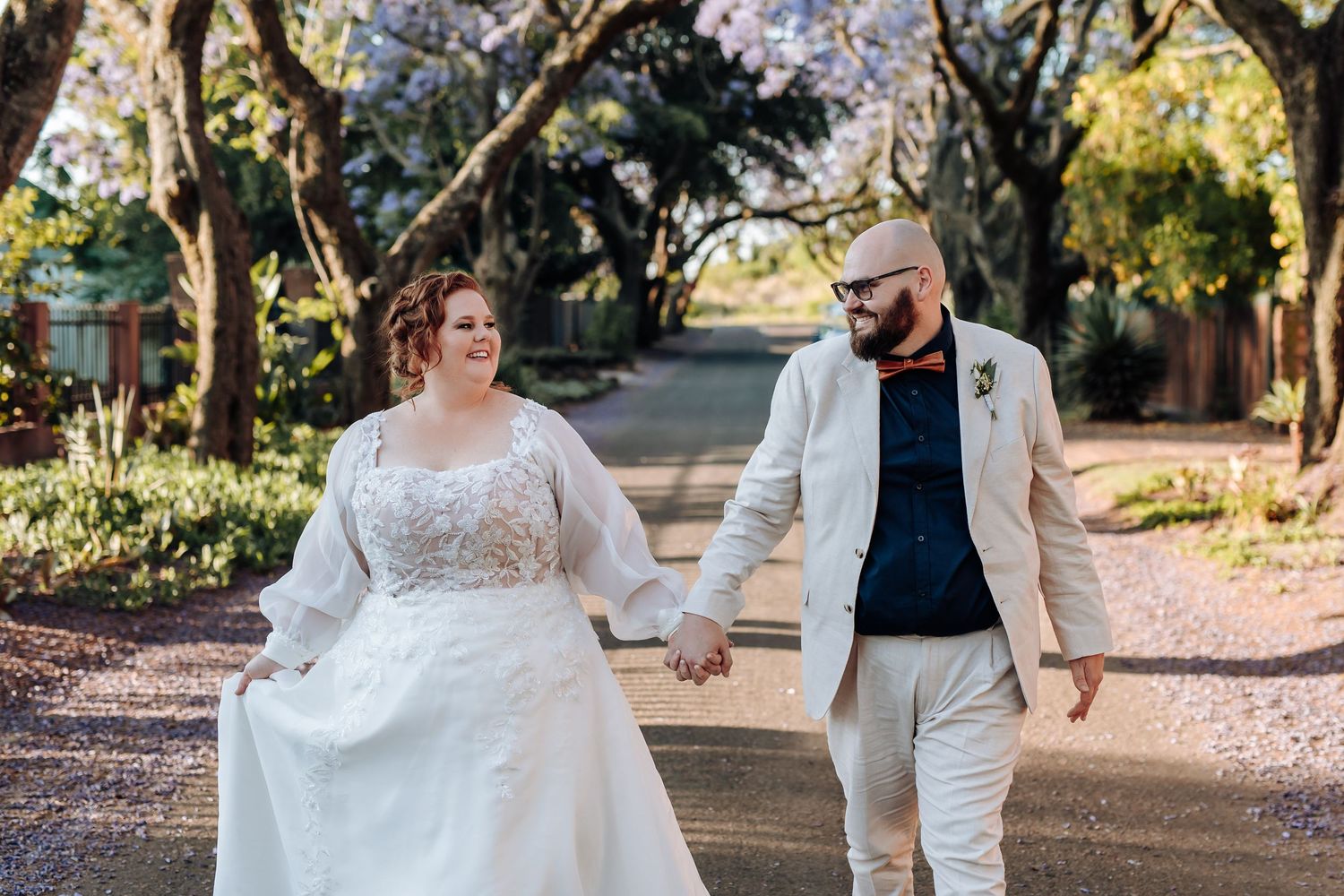 A couple in wedding attire walks hand in hand down a tree-lined path on their special day.