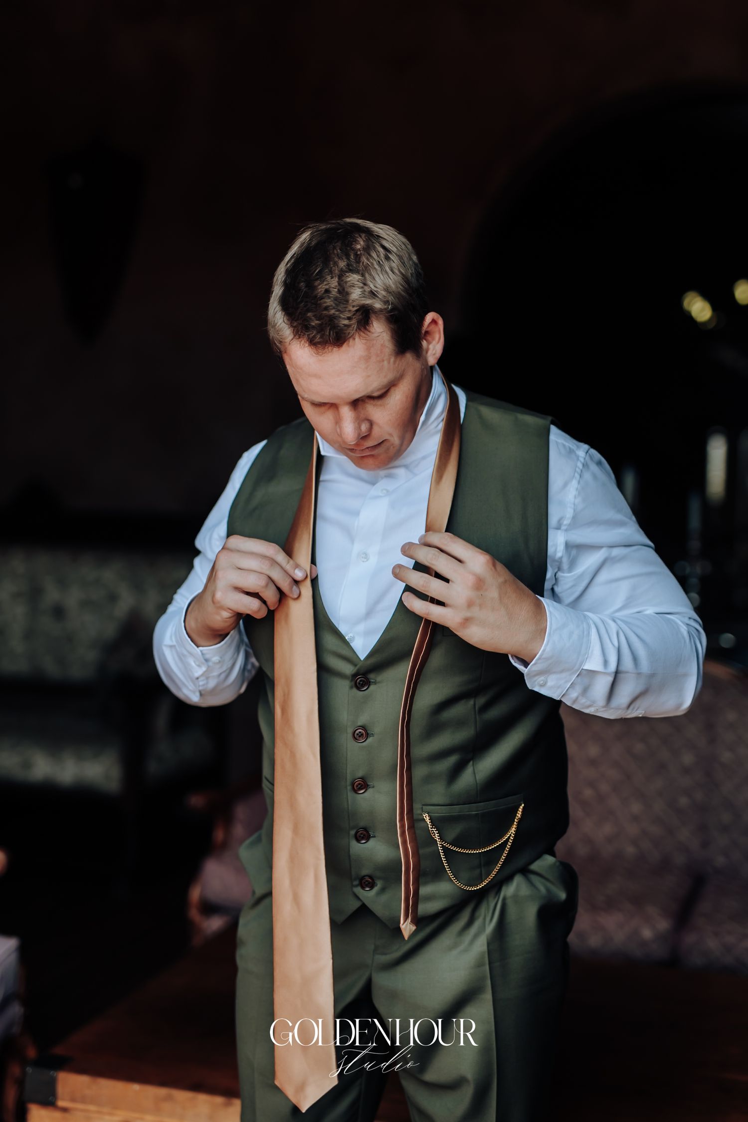 Groom in green vest adjusting gold tie with pocket watch chain detail.