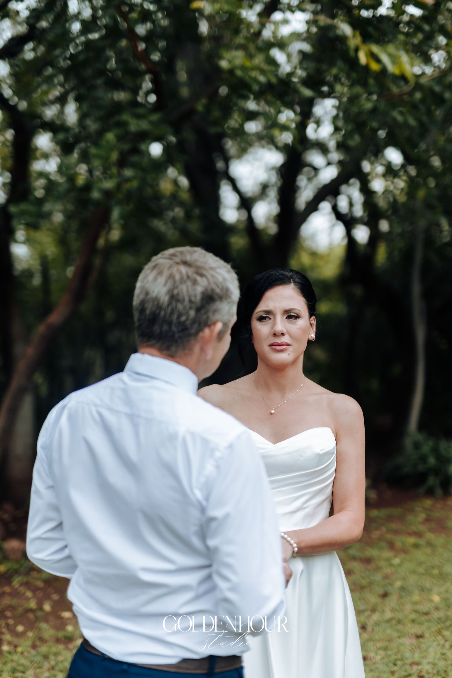 Bride in strapless white gown holding hands with man in white shirt during outdoor wedding ceremony.