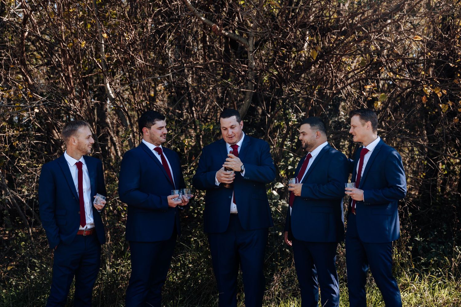 Five groomsmen in navy suits and red ties celebrate outdoors with drinks before a wedding ceremony.