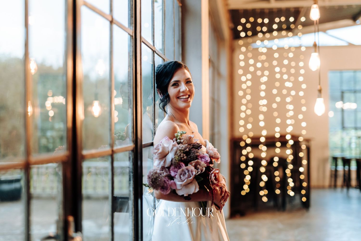 Elegant bride in strapless gown holding a pink and burgundy bouquet, smiling beside large windows with fairy lights.