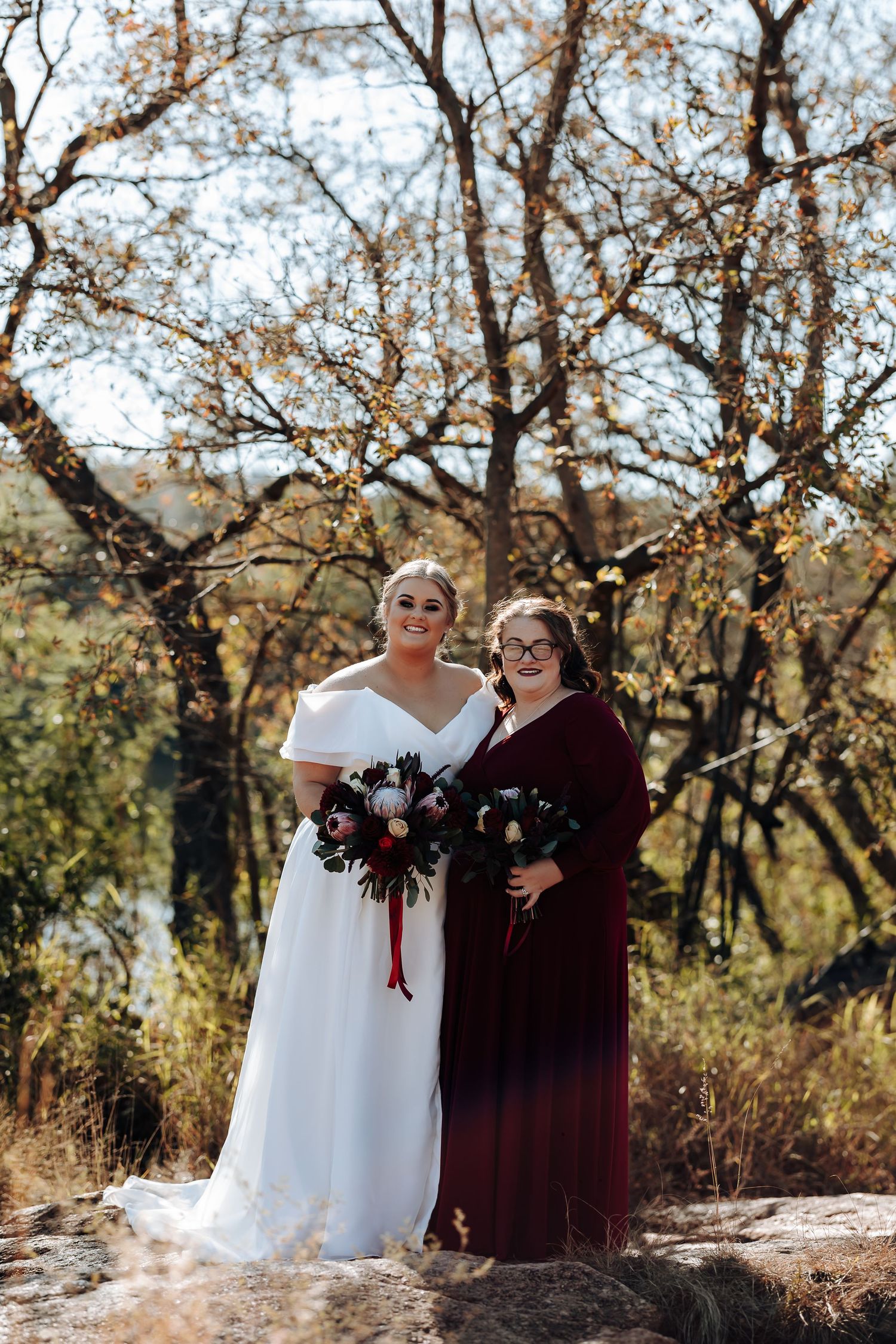 Bride in white gown and bridesmaid in burgundy dress holding bouquets outdoors in autumn setting.