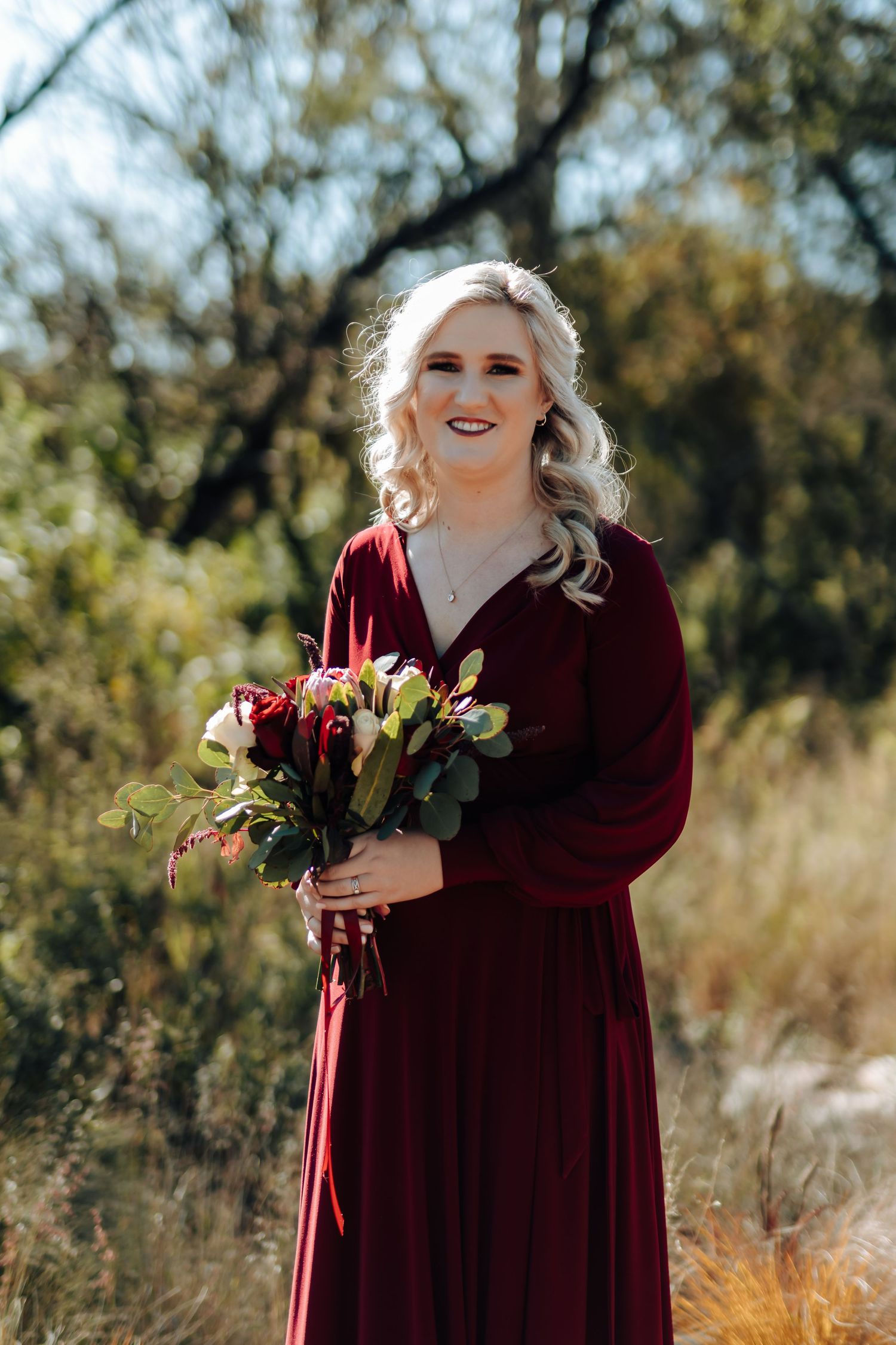 Woman in burgundy gown holding floral bouquet smiling outdoors with natural greenery background.