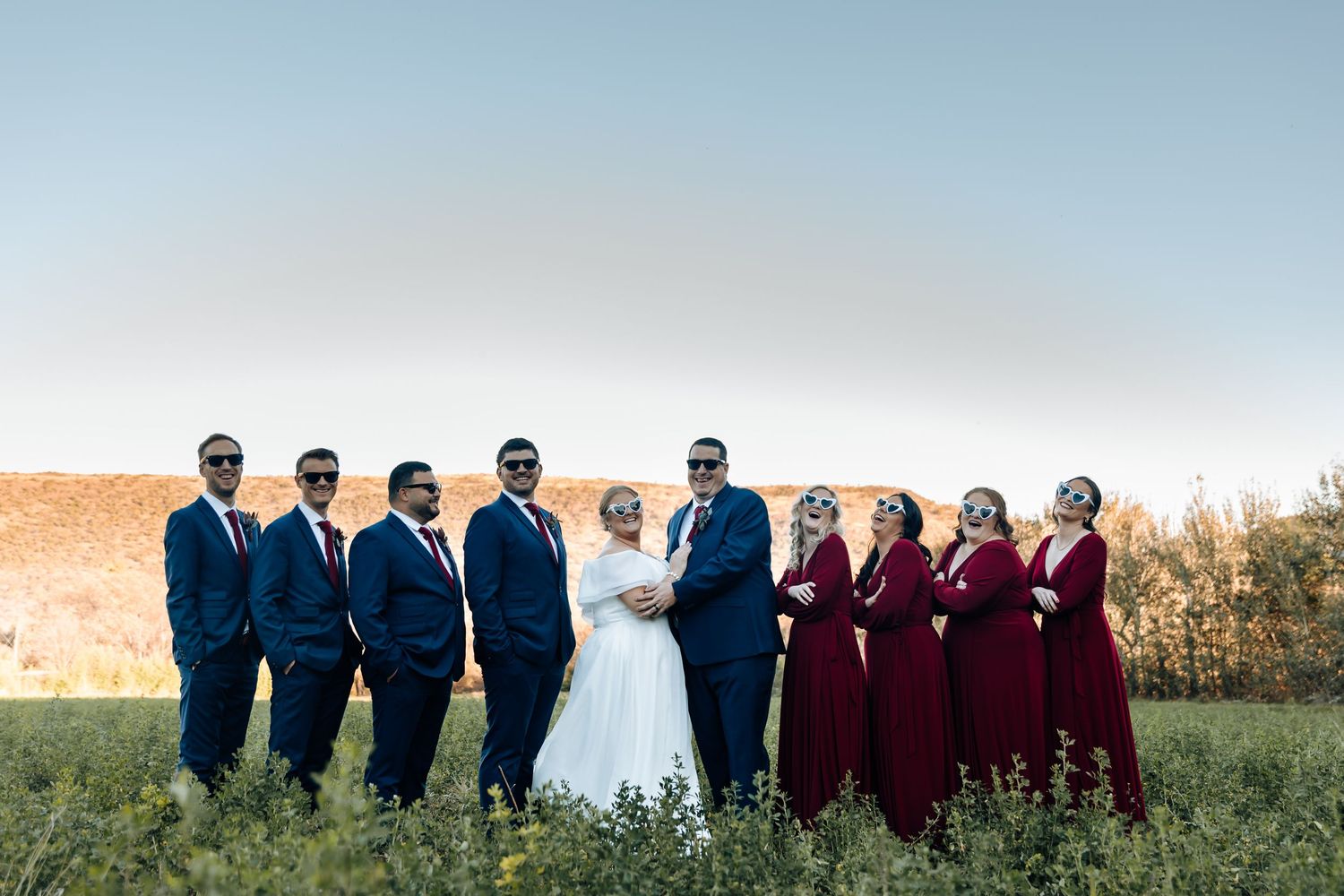 Wedding party posing outdoors, groomsmen in navy suits with sunglasses and bridesmaids in burgundy gowns flanking the bride and groom.