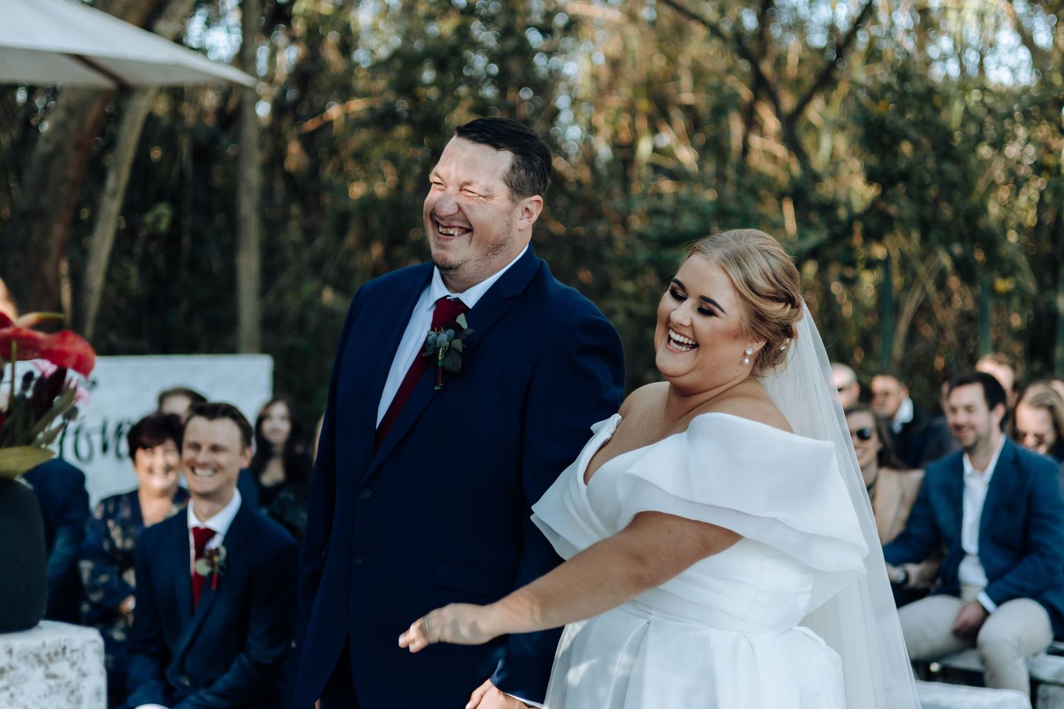 Joyful bride in off-shoulder white gown laughing with groom in navy suit during outdoor wedding ceremony.