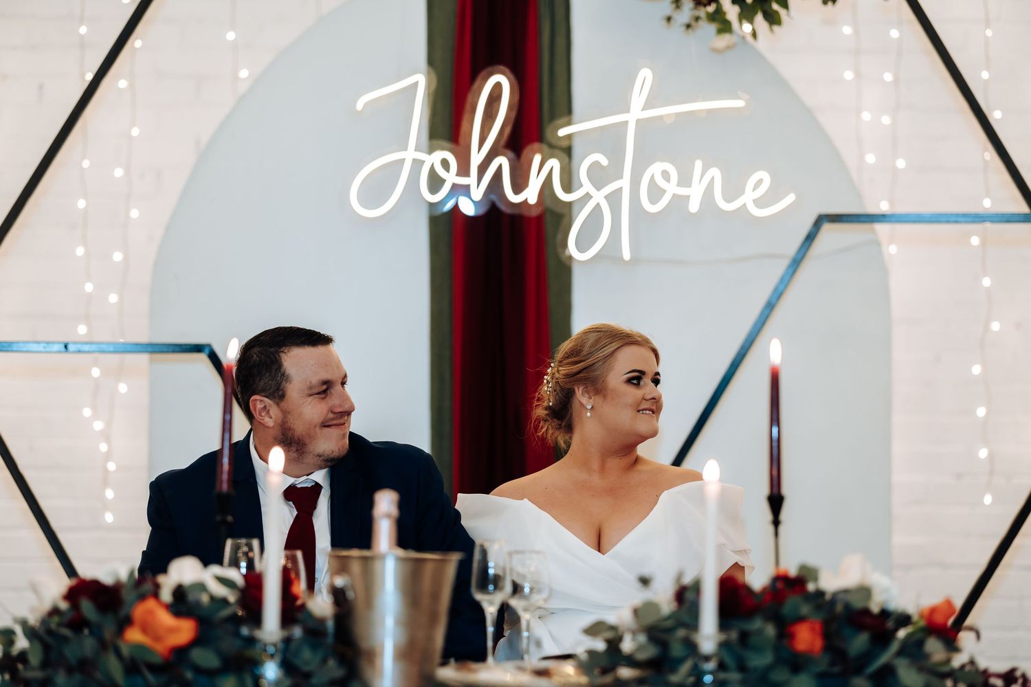 Bride and groom smiling at wedding reception under Johnstone neon sign with fairy lights and floral centerpieces.