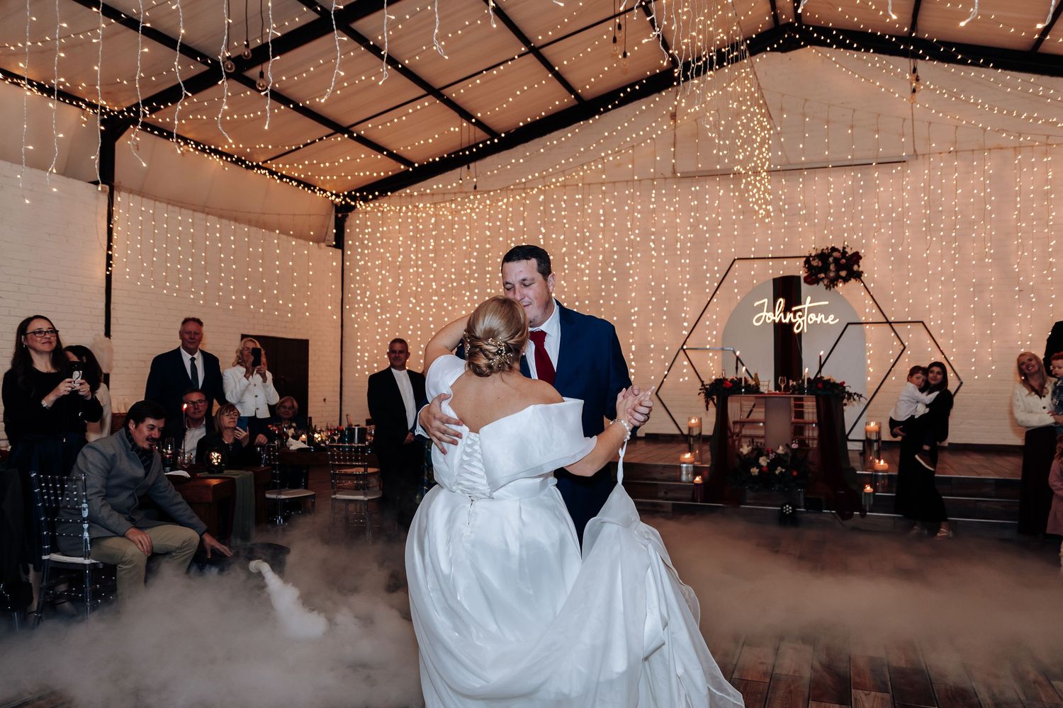 Bride and groom share romantic first dance on fog-filled dance floor under twinkling fairy lights at elegant wedding reception.